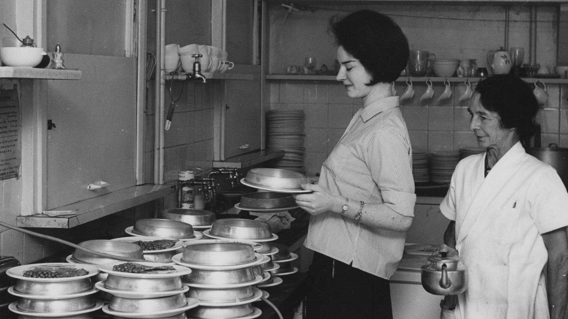 Black and white photo shows two women in a kitchen, one of whom is holding a kettle and wearing a white jacket. In front of them, there are dozens of dishes with peas covered by metal hot plates