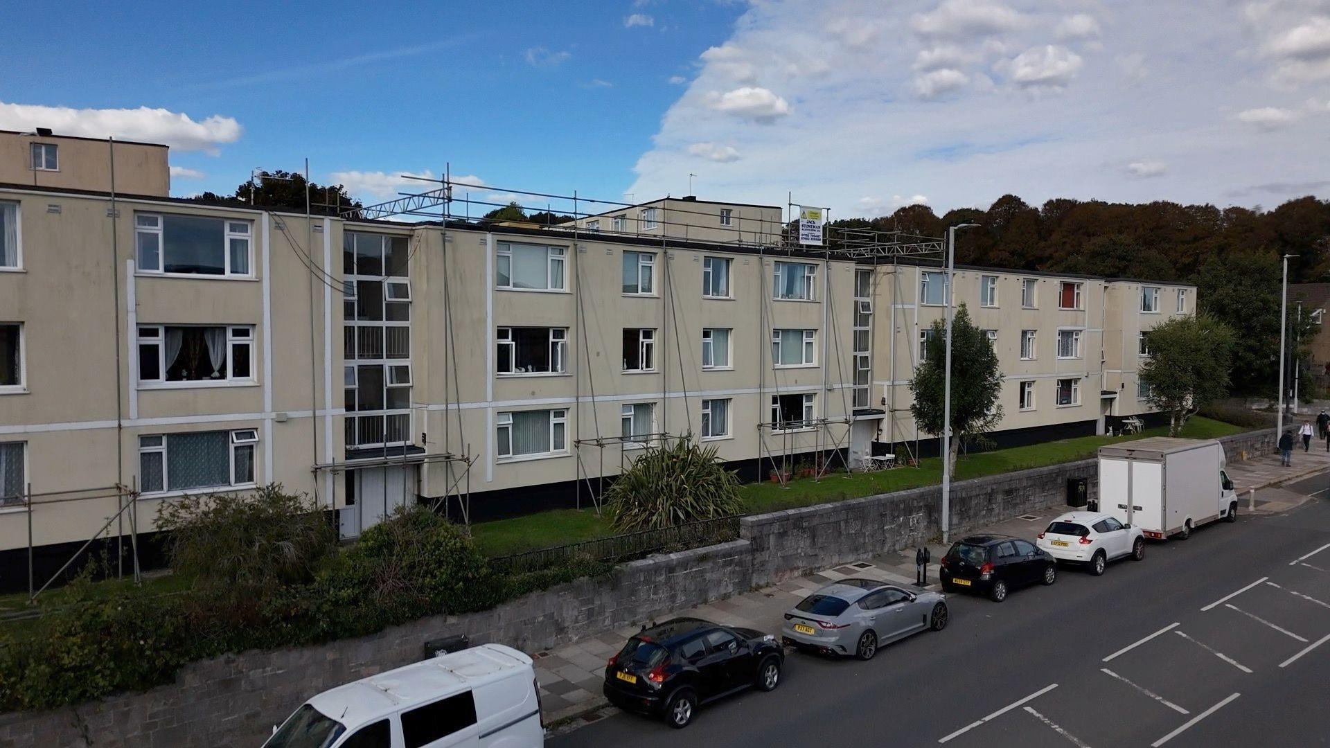 A cream coloured three-storey block of flats with a scaffolding tower and further scaffolding to the right of the picture. Only part of the block and the top two storeys are visible.