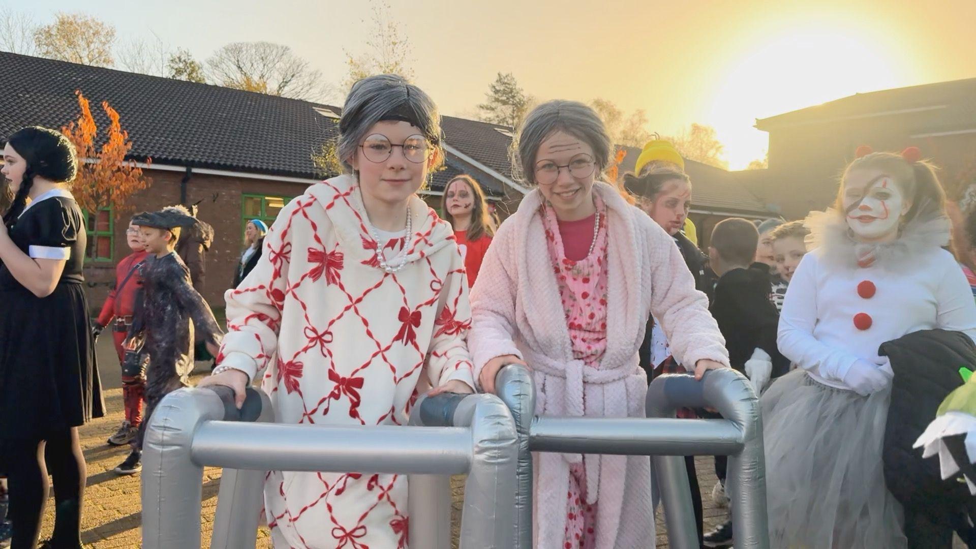 Two girls dressed as grannies - they have grey wigs on and are both wearing dressing gowns, the girl on the right's is pink and her friend's is white with a red bow pattern on it. They are both holding inflatable zimmer frames.