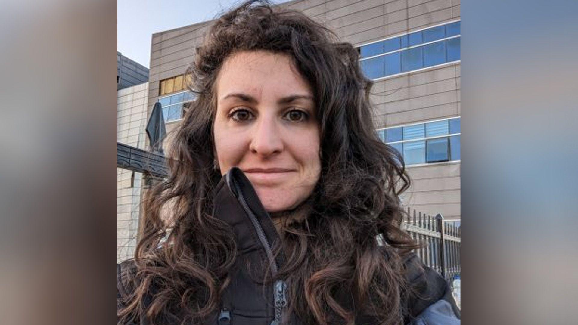 Ms Morelli wearing a black jacket looks at the camera in front of a building and a black railing fence. 