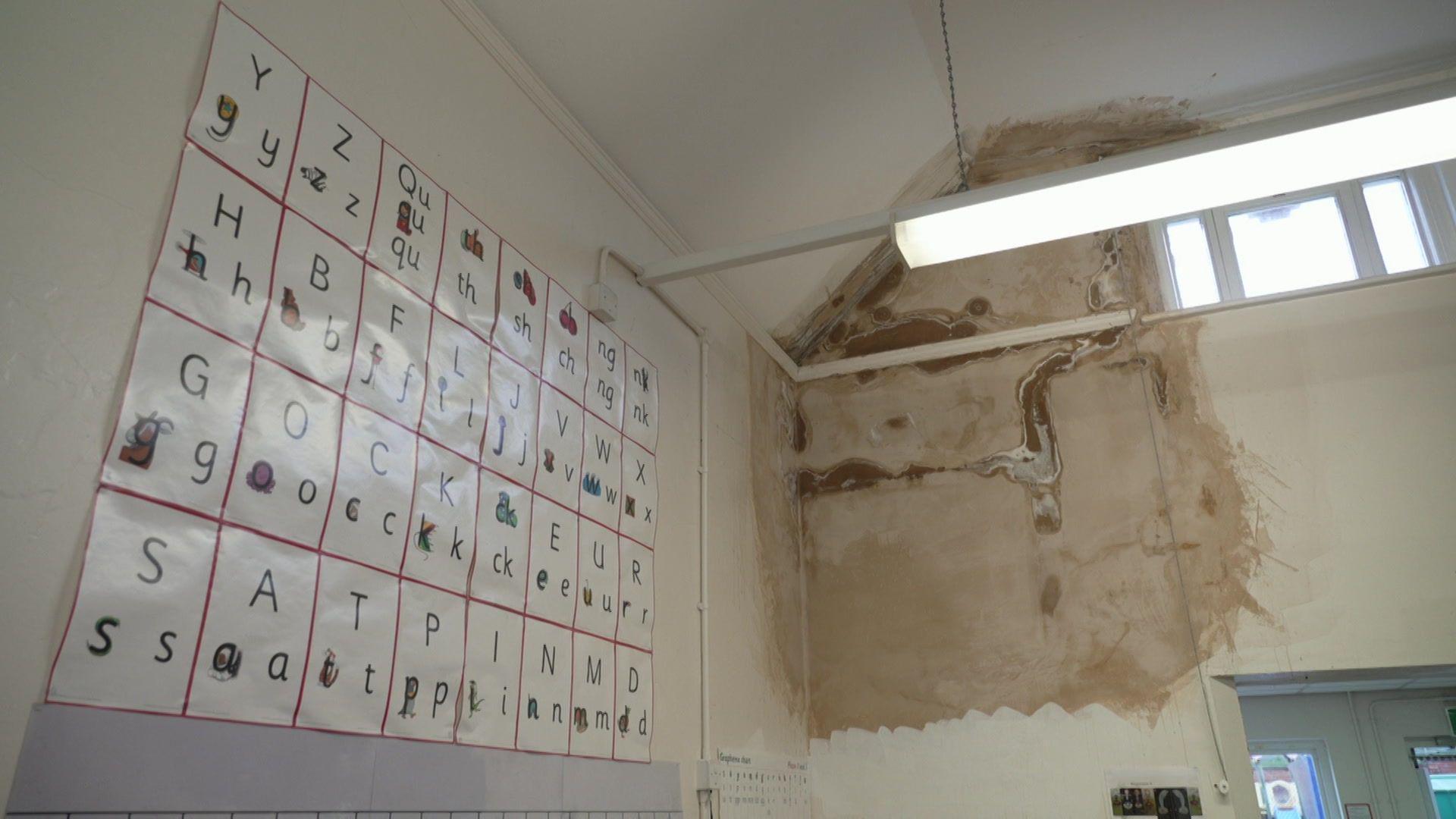 A classroom with an alphabet board on one wall. The wall next to it has a large, dirty, brown damp patch leading from the roof, where a leak has been