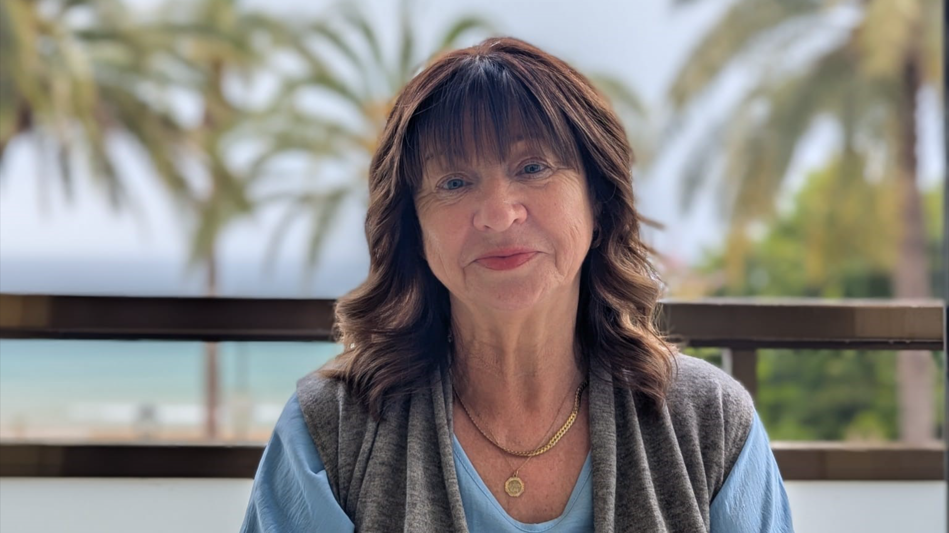 Kathleen Richards, who has dark shoulder-length hair with a fringe, is sitting on a balcony in front of the sea. She is wearing a blue top and a grey, sleeveless cardigan. Palm trees can be seen in the background.