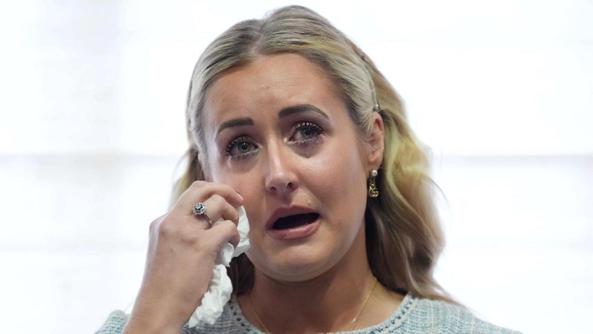 Katie Amess uses a tissue to wipe away tears during a press conference. She has blonde hair and is wearing a knitted blue cardigan.