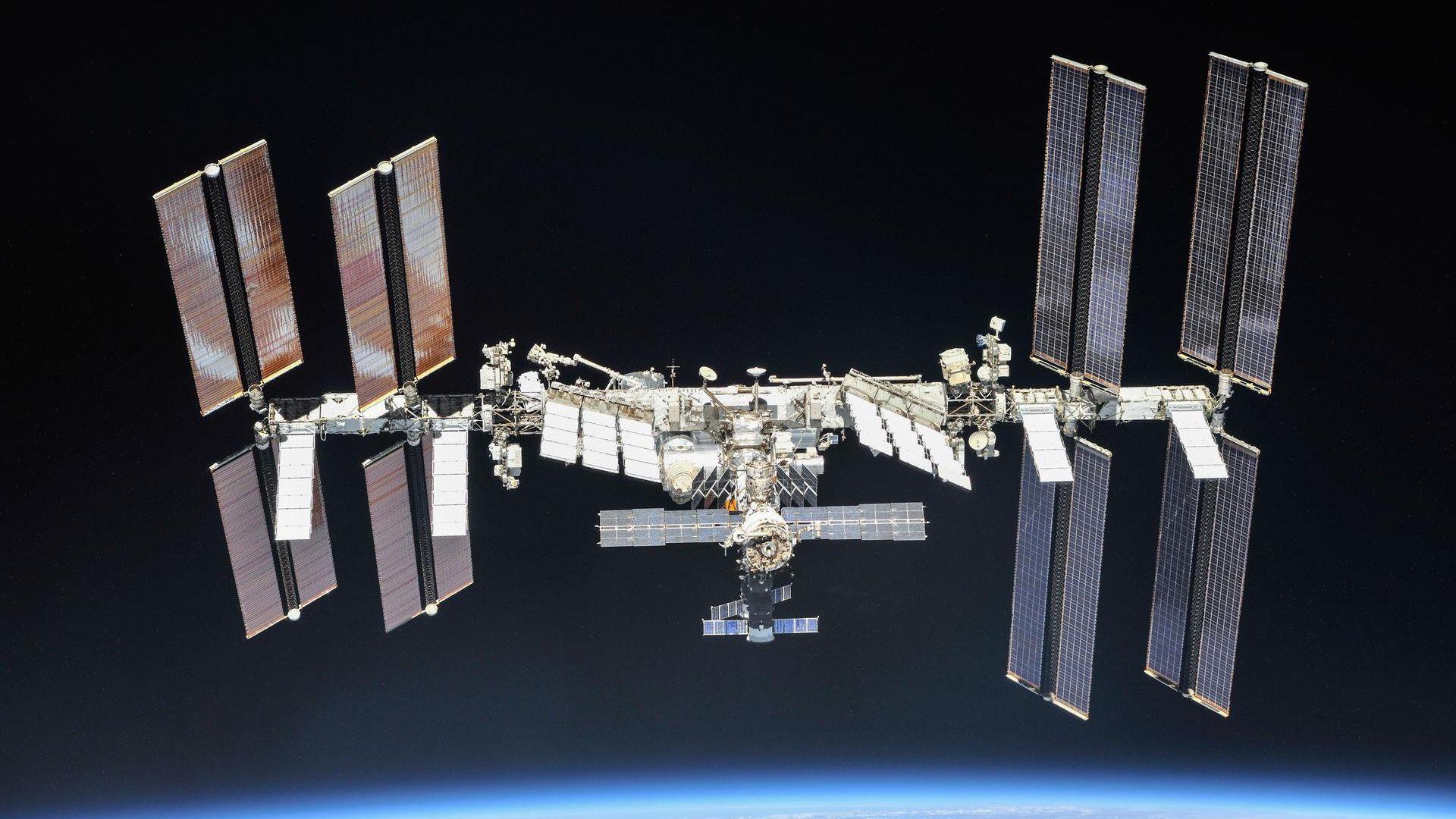 A picture of the International Space Station with big panels coming out of it. It's surrounded by darkness and there's a faint blue glow from the Earth at the bottom of the picture