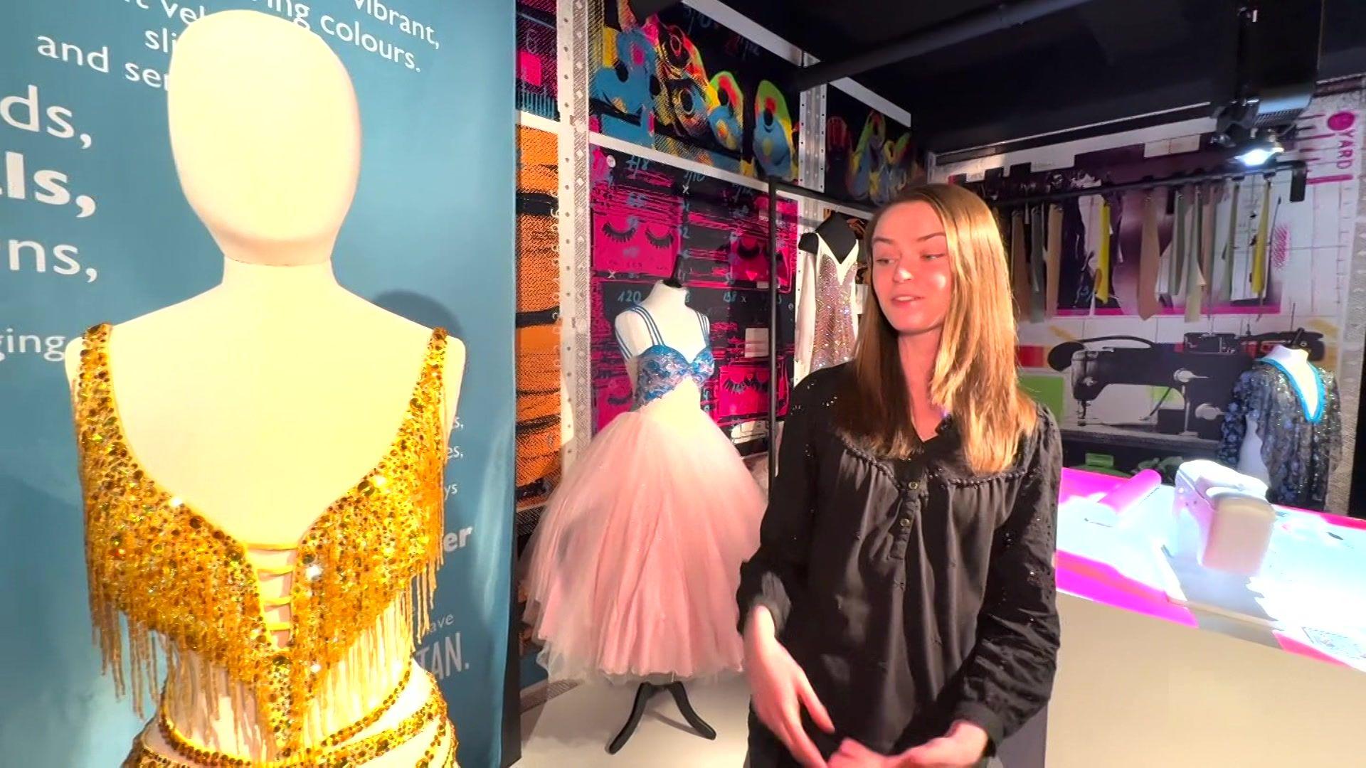 Showtown exhibitions assistant, Emily Langridge wears a black blouse and has long blonde hair. She is standing next to a mannequin which has Anita Rani's tassled sunrise yellow dress, which has topaz crystals. It is a strappy cut out short dress.