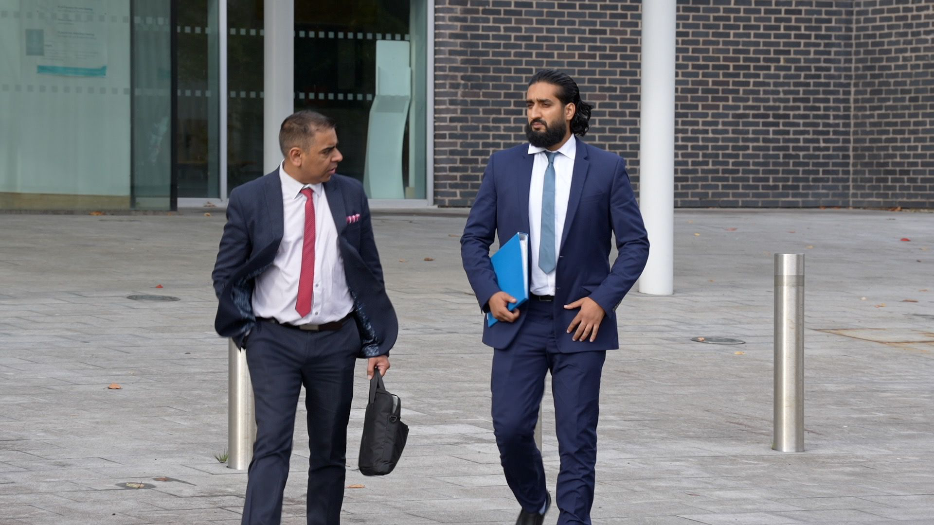 Photograph showing Abubakr Ben Yusaf, who wears a navy two piece suit, a blue tie and white shirt. He has brown long hair and a brown long haired beard. He is leaving Mold Crown Court and holds a blue folder under his left arm.