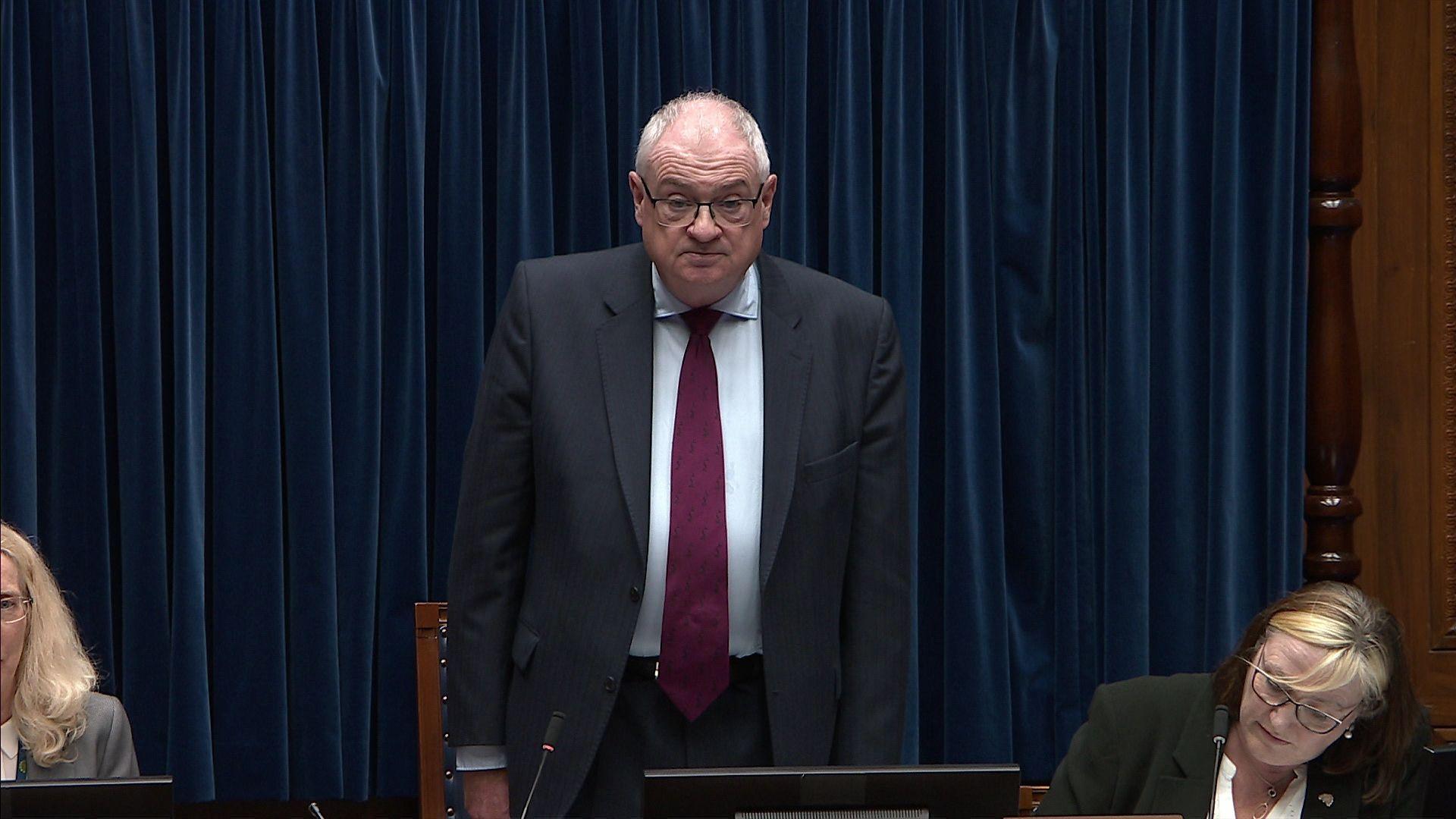 Steve Aiken stood against a blue curtain in NI assembly, he has short grey hair and is wearing a navy suit, white shirt and purple tie