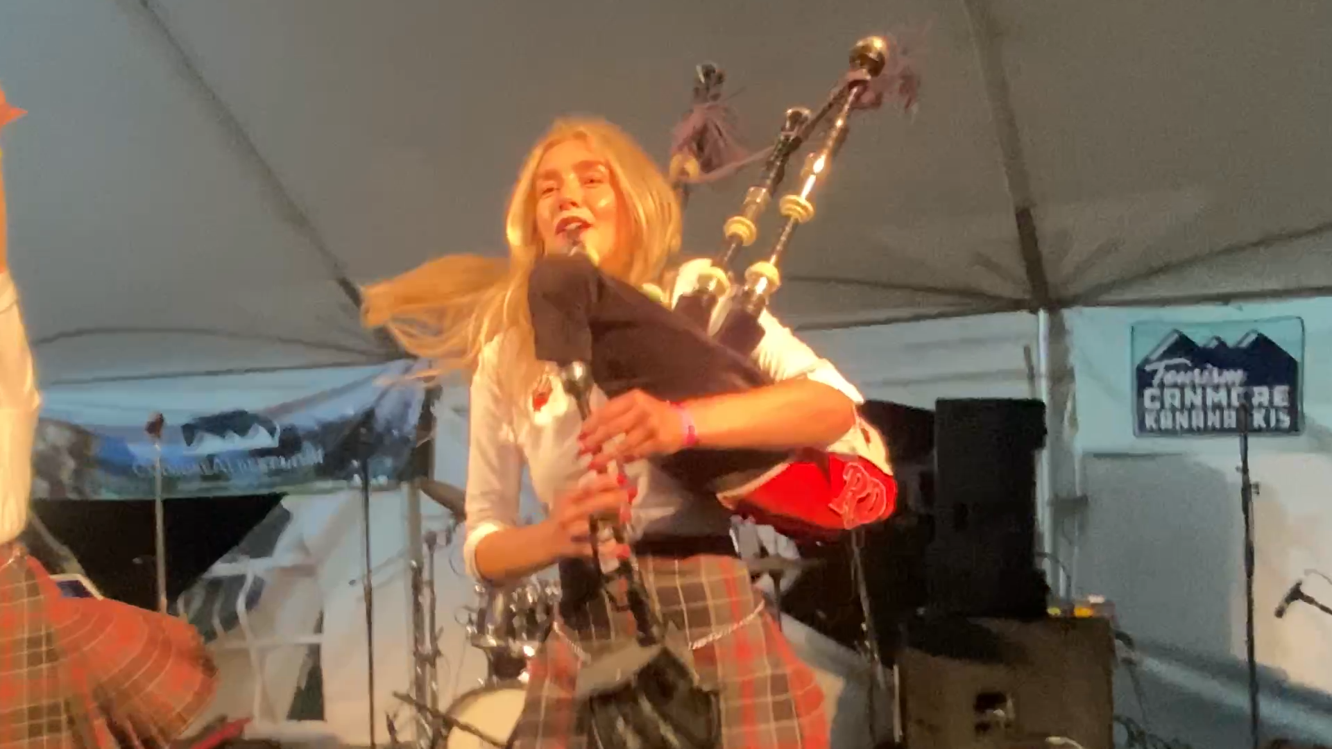 Bagpiper Anna Smart on stage playing bagpipes in a brown and red kilt. She has long blonde hair and a brown set of bagpipes.