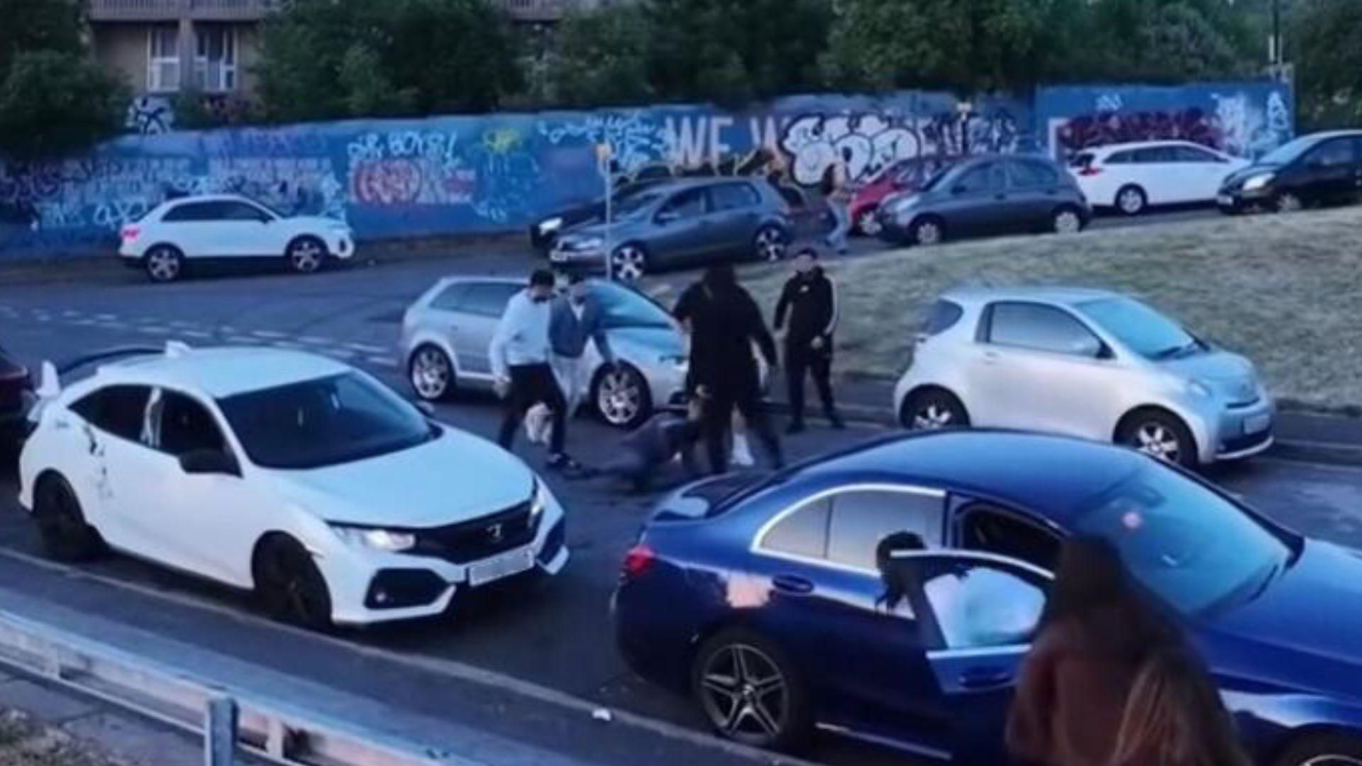 Many cars parked around and five people kicking a man on the floor in the road
