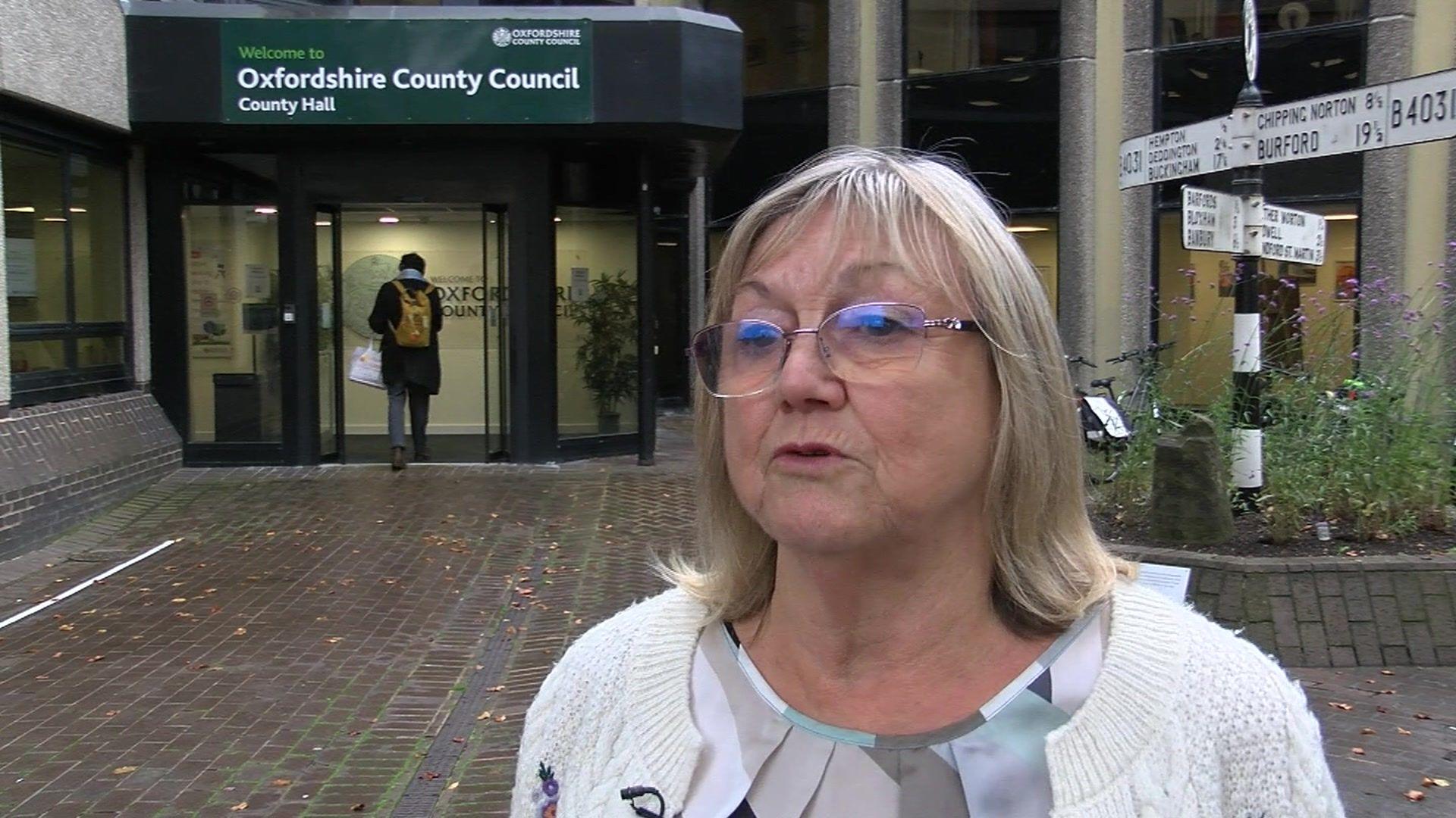 Judy Roberts has medium length grey hair, glasses and is wearing a beige cardigan. Behind her is the front of Oxfordshire County Council's building.