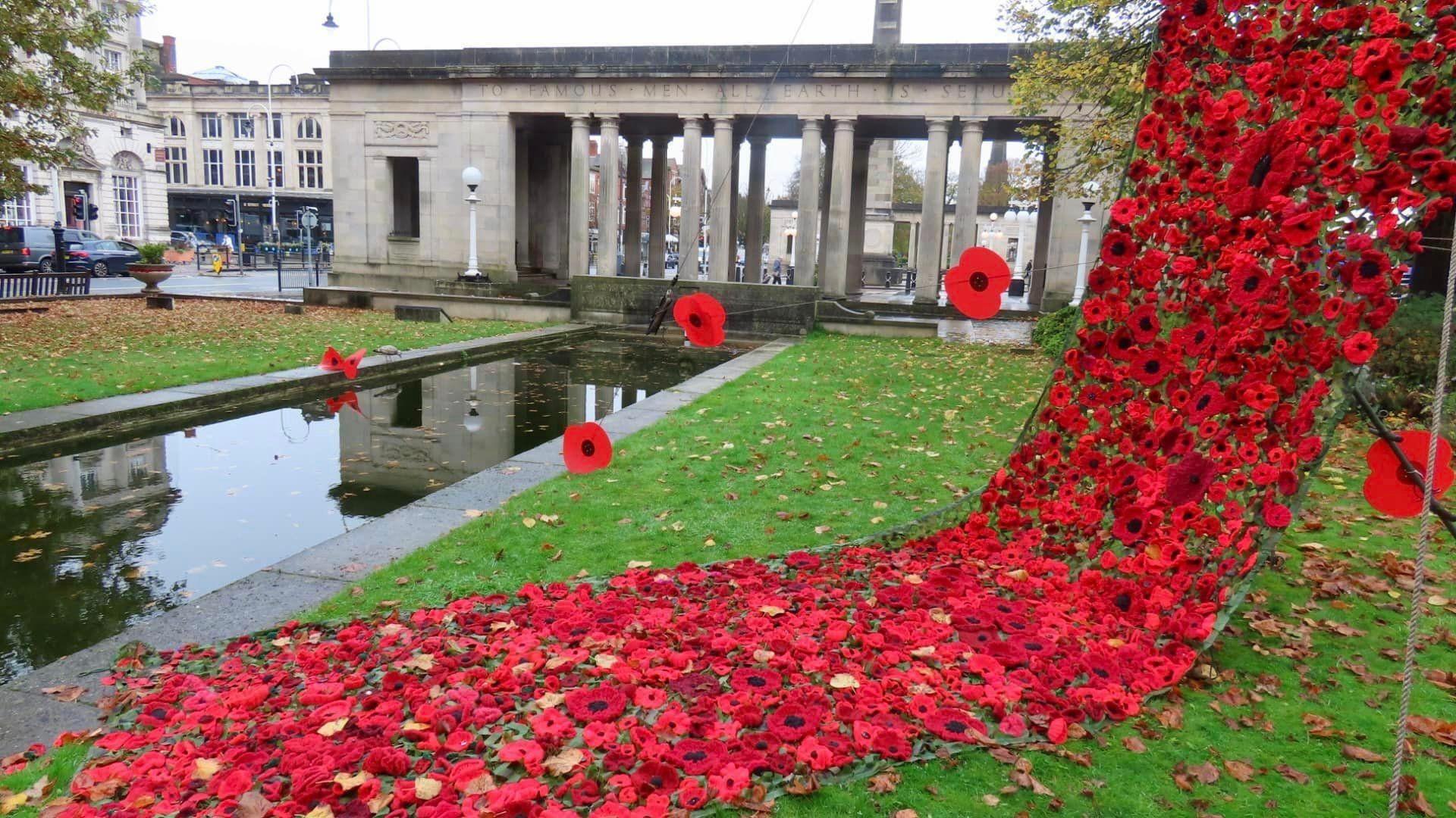 Red knitted poppies hang down in a drape, with purple knitted poppies forming one giant poppy within the drape. It hangs down in Remembrance gardens in Southport, on an autumn day