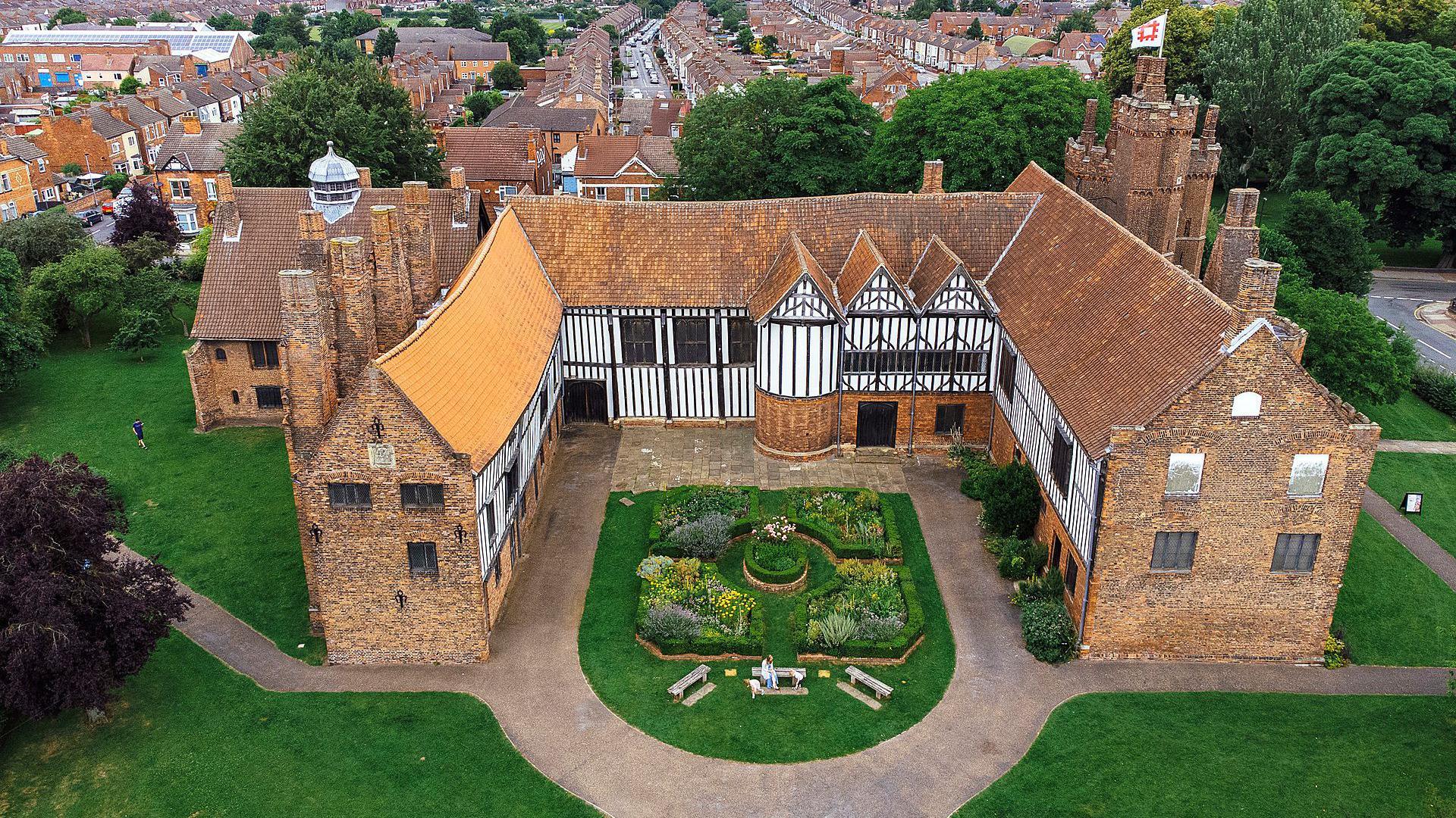 A medieval manor house with the photo taken of it from up in the sky - it looks like it has been taken from a drone. Distinctive black and white markings can be seen on its sides with a patch of greenery in between the walls. Greenery stretches around the manor with terraced houses seen in the distance.