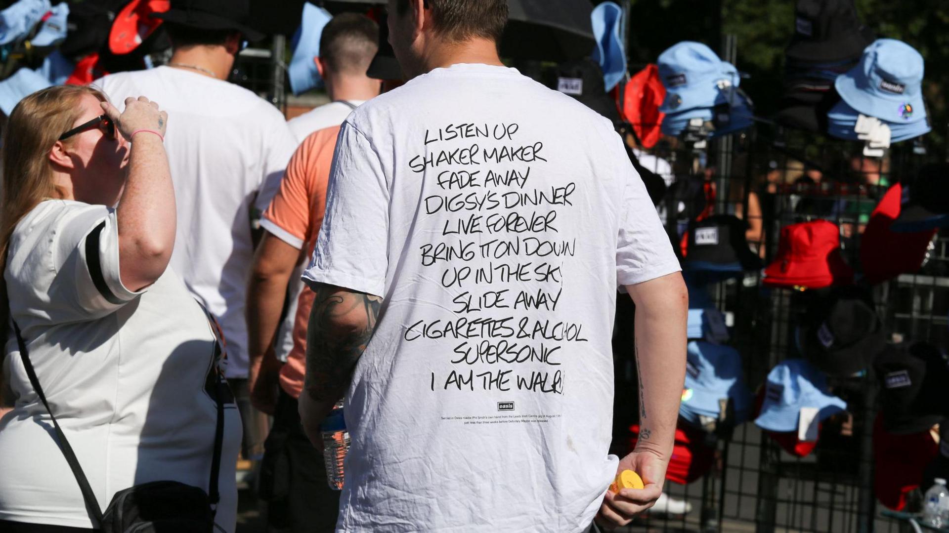 A man in a white T-shirt stands in front of a stall selling red and blue bucket hats. Words on the T-shirt read: Listen Up, Shakermaker, Fade Away, Digsy's Dinner, Live Forever, Bring It On Down, Up In The Sky, Slide Away, Cigarettes & Alcohol, Supersonic, I Am The Walrus