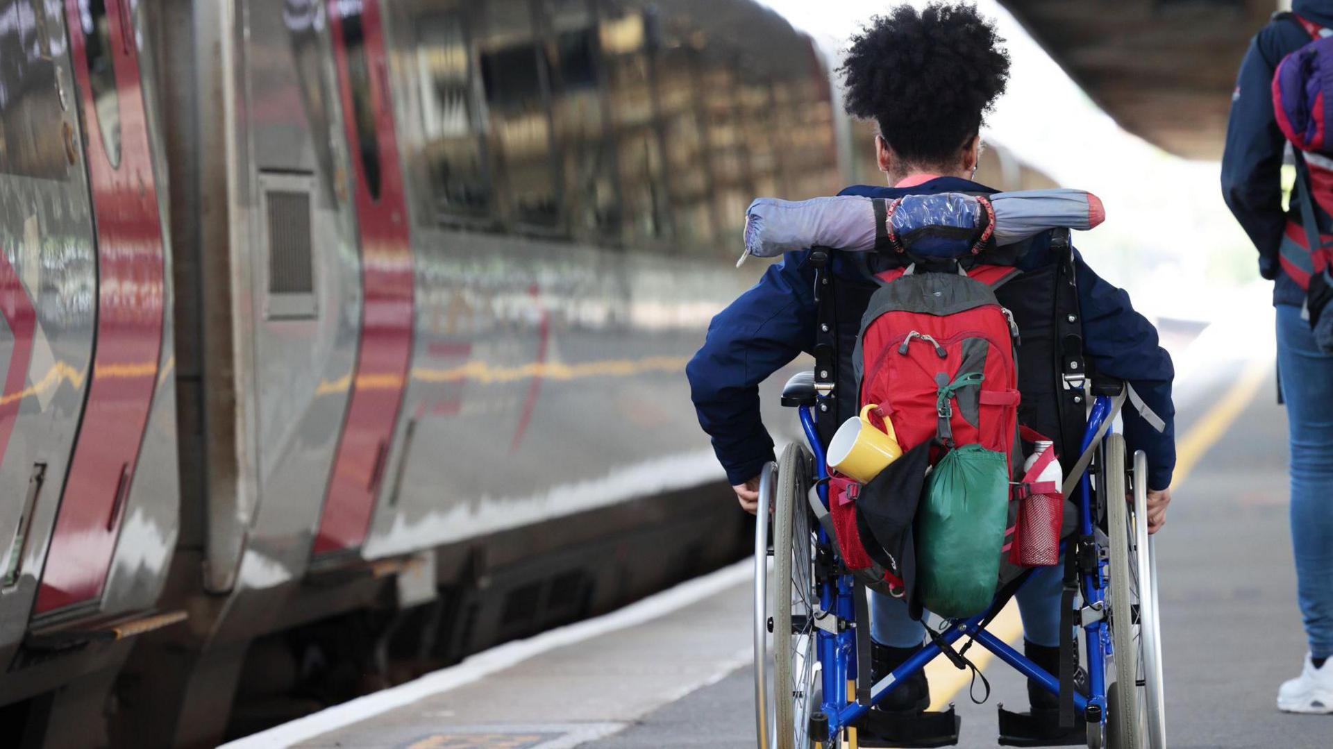 A woman in a wheelchair is visible from behind, wheeling herself along a train platform. She is wearing a rucksack and has a blue jacket on. She has black hair. There is a train on the platform to her left, with a grey and pink livery.