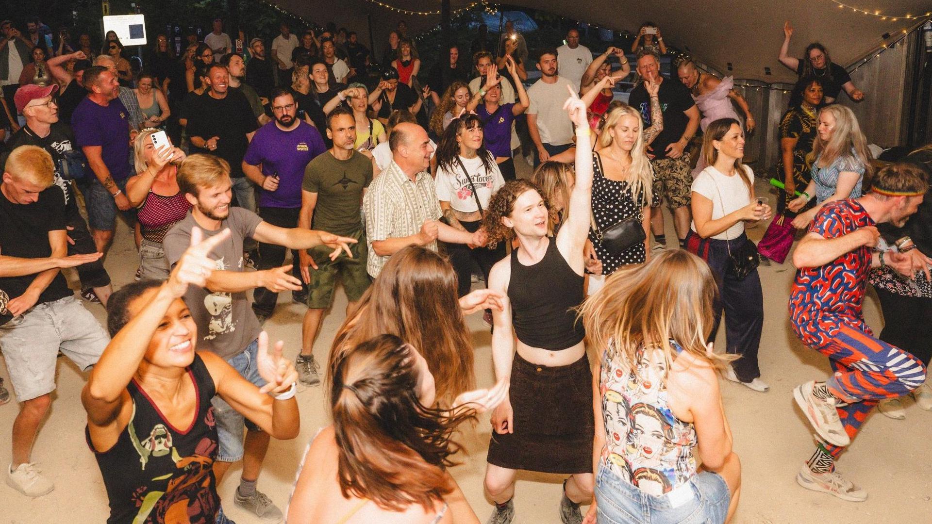 A group of people dancing underneath a marquee. It is dark outside and there are strings of lights draped across the top of the marquee.