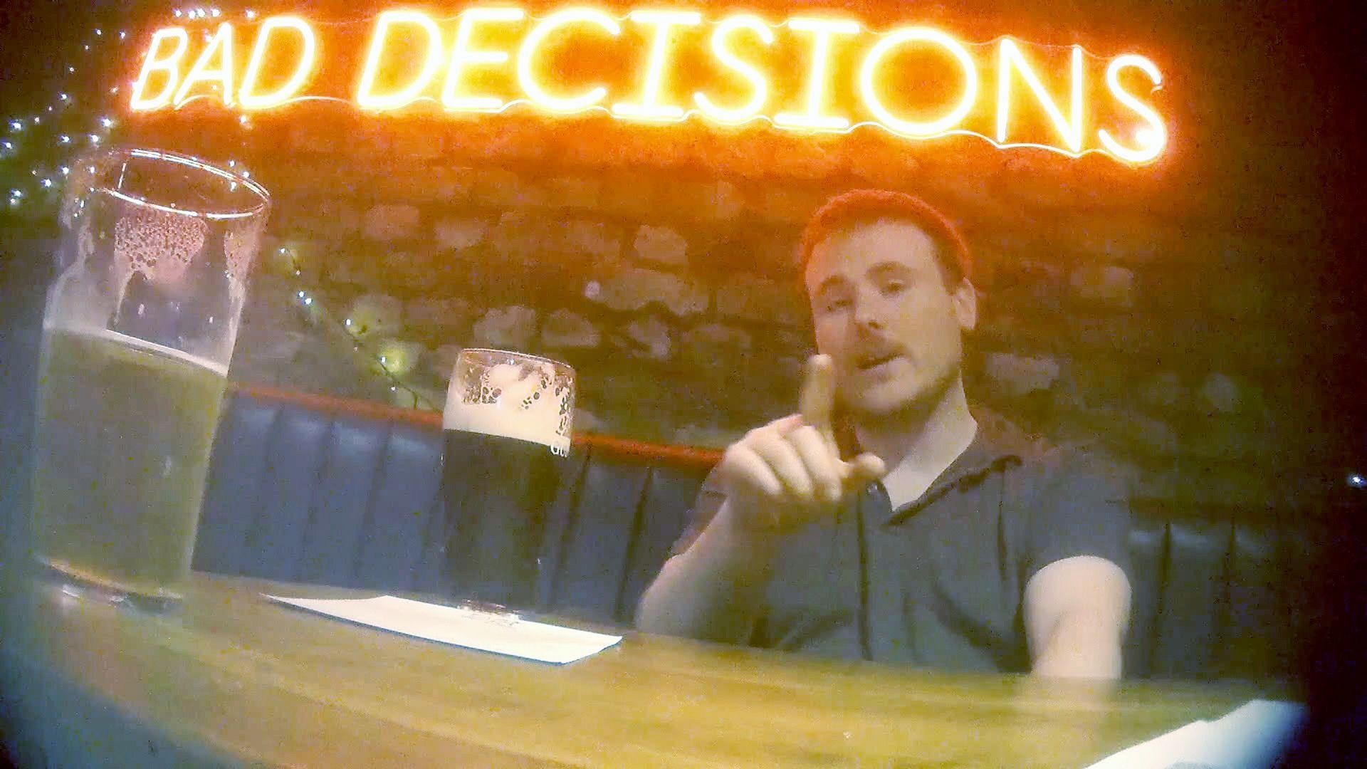 A hidden camera shot of PC Phil Neilson taken from across a pub table. PC Neilson wears a black polo shirt and had grown a moustache at the time; he is pointing at Rory across the table as he talks and drinks a Guinness. Above his head on the bare brick wall is a red neon sign which reads: "BAD DECISIONS".
