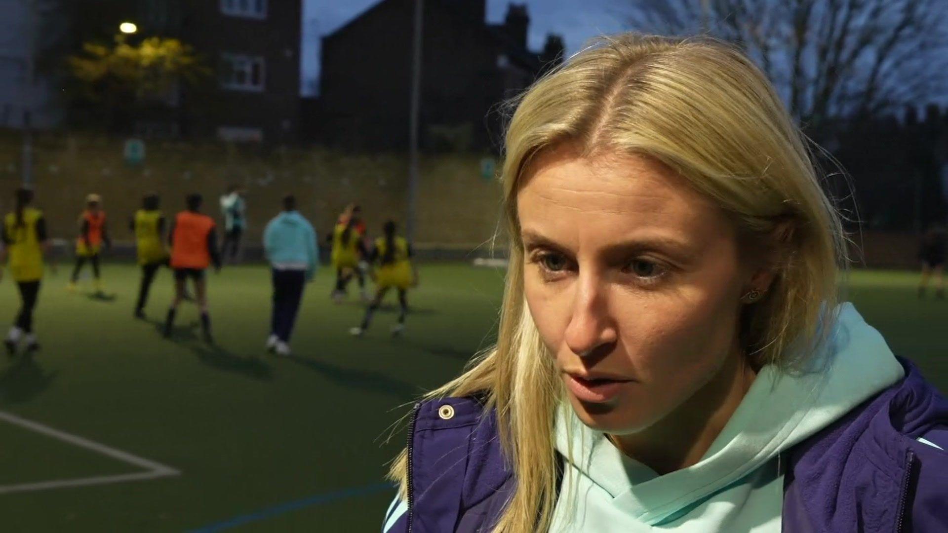 TV screenshot of Leah Williamson, a footballer. She is standing by the edge of a football pitch on which a training session is taking place. It is dark and the pitch's lights are on. Leah is wearing a pale blue hoodie and a purple coat