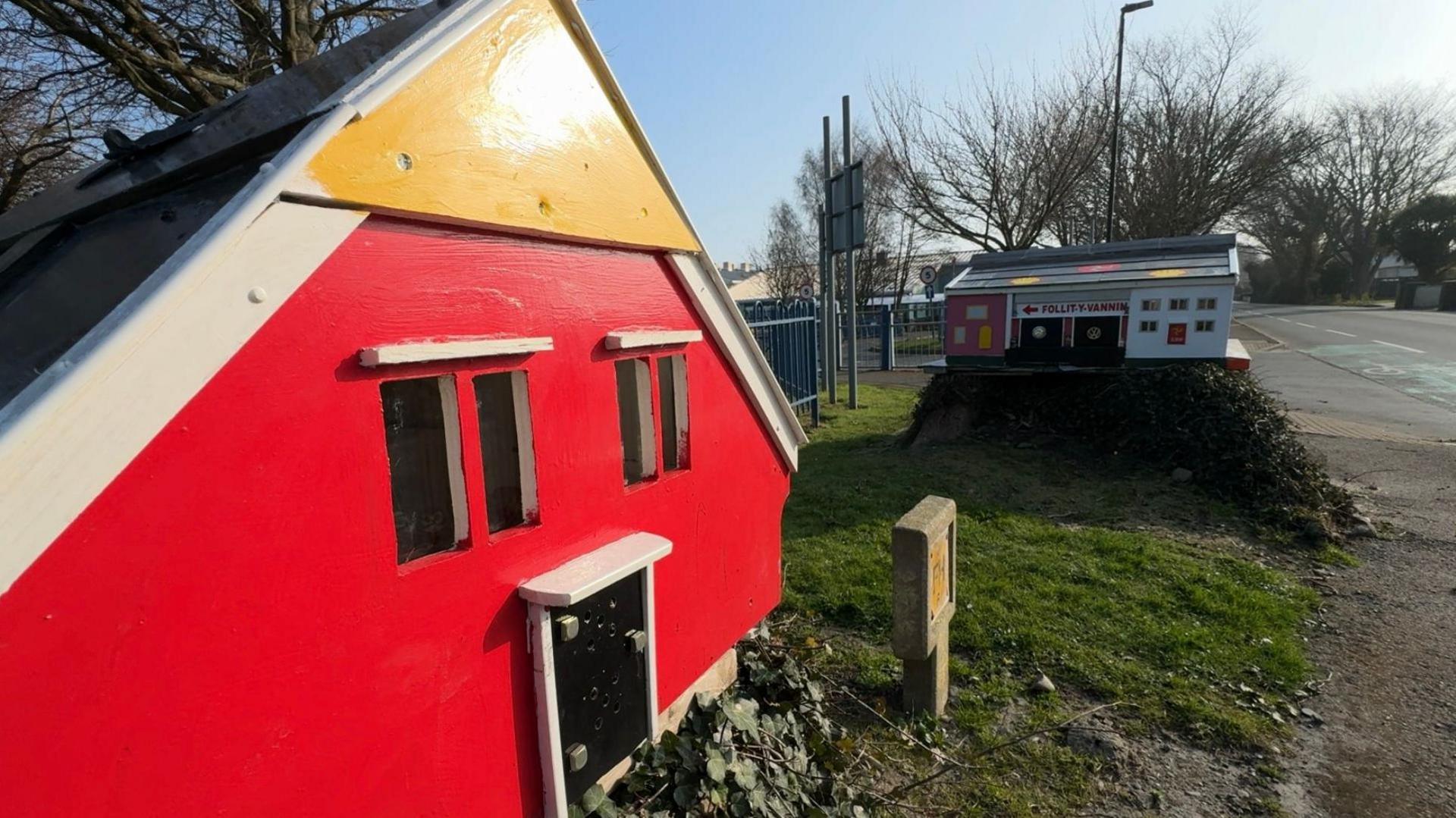 A red fairy house with black doors and windows can be seen in the foreground, the new house can been seen in the distance on a tree stump.