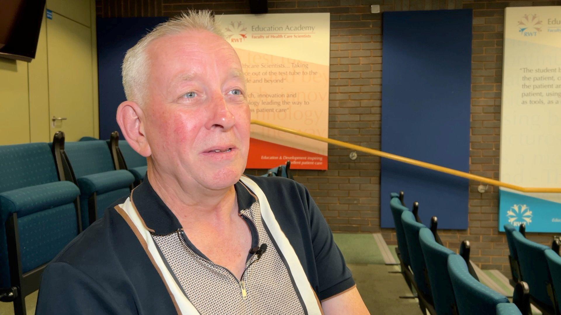 Wayne Devaney, he is wearing a polo shirt with a zip collar. He is sitting in a room with a yellow hand rail, chairs and signs visible in the background