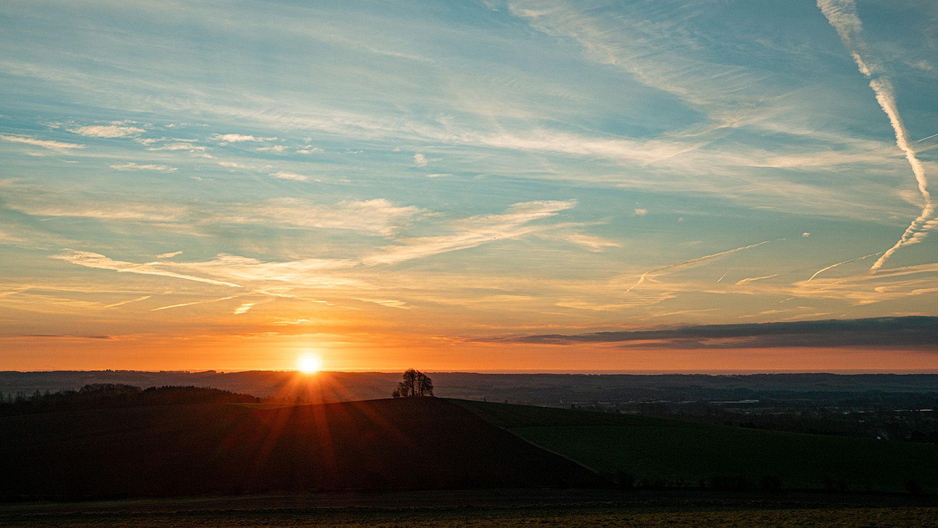 The sun appears over the horizon behind rolling hills. A group of trees is on the brow of the hill. The sky above is orange and blue with streaks of cloud.