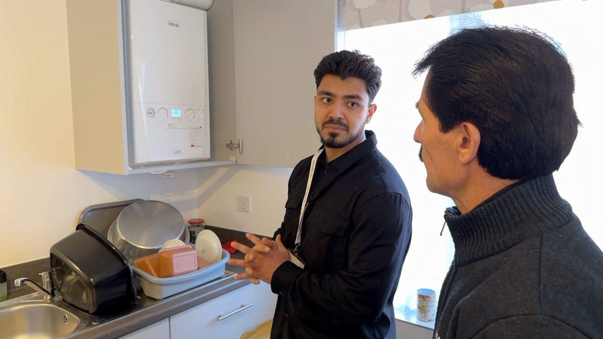 Two men stand in a small kitchen next to a sink with washing up on the draining board and a bowl to one side. One of the men wears a black shirt with a lanyard around his neck and has his hands clasped, while the other man wears a turtleneck jumper and is looking at him.