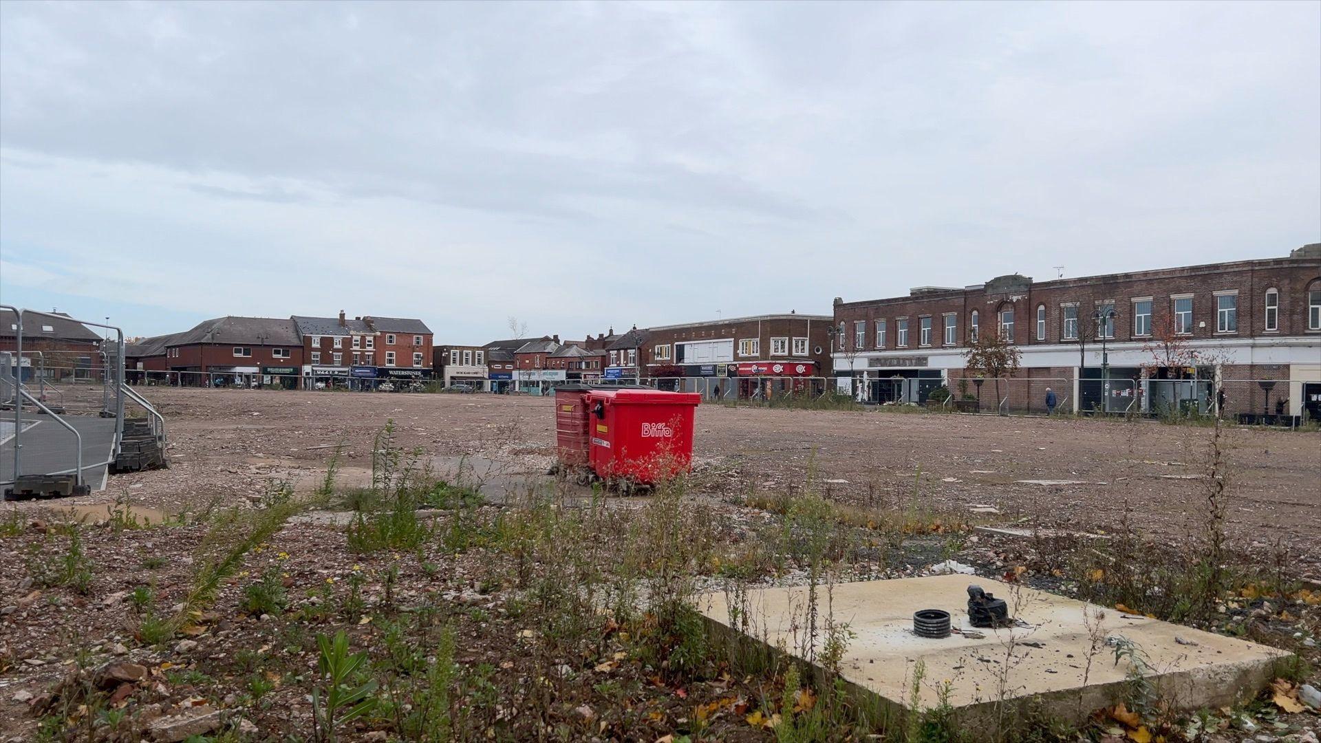 A view of the empty site in Crewe town centre that has been saved for the second phase of the Royal Arcade development. It is flat, undeveloped land with weeds poking out, surrounded by old brick buildings in the background