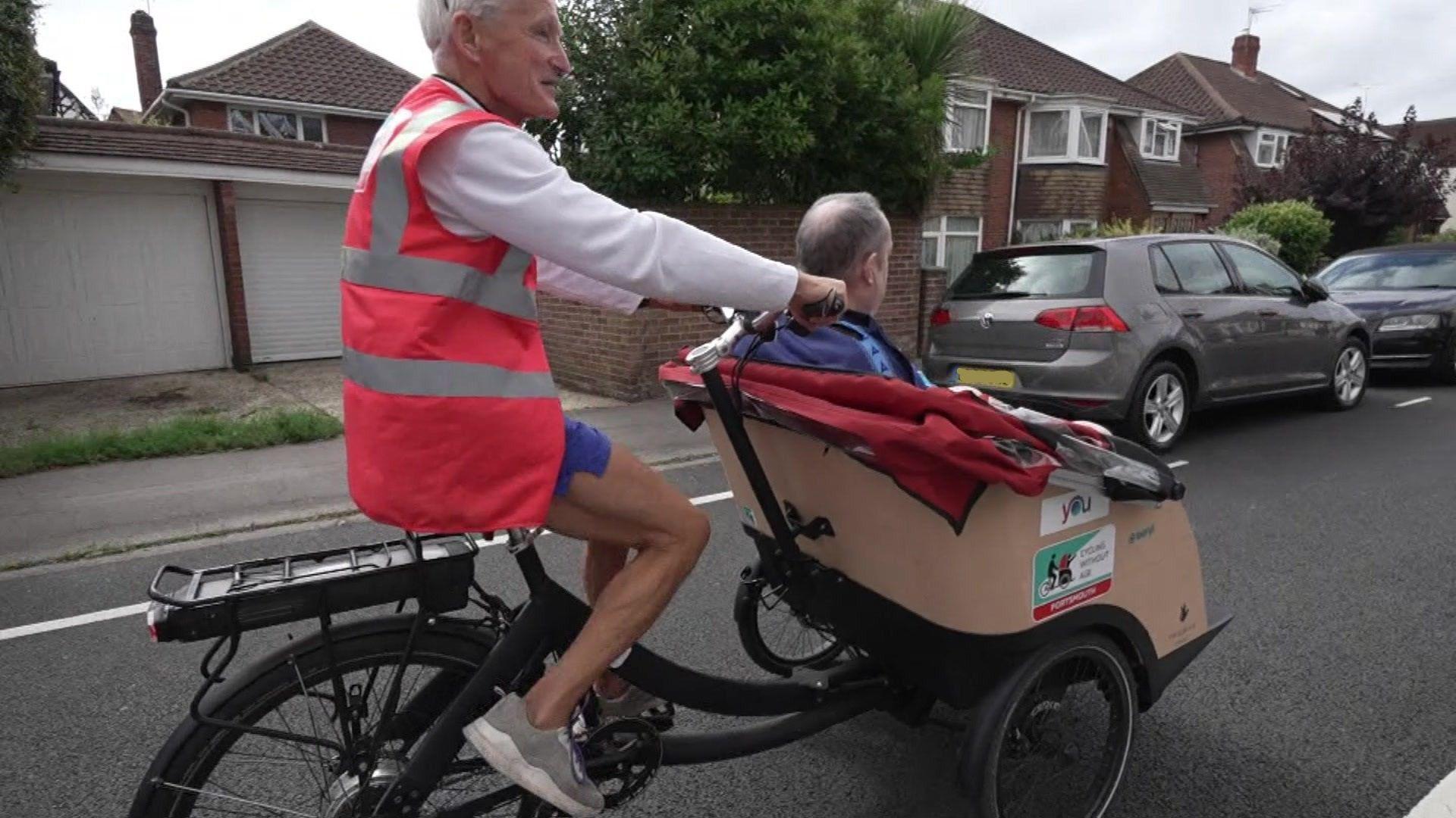 A man with grey hair and wearing a red high-visibility jacket cycles down a residential street on a bike with a passenger cart fixed to the front. Inside the cart, a man in a blue top with grey hair is seated, facing away from the camera.