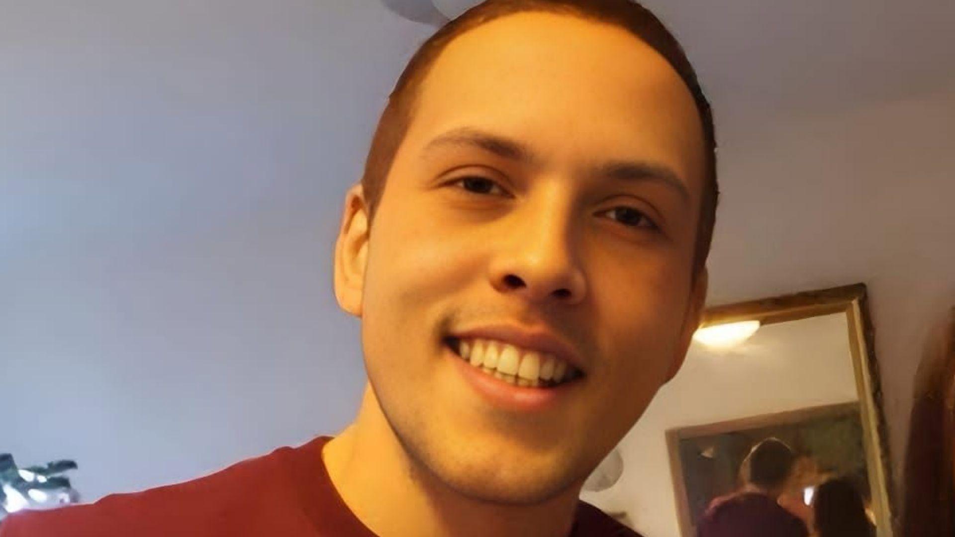 Close-up photograph of Gareth looking towards the camera indoors, with a mirror visible in the background. He has cropped brown hair and is smiling.