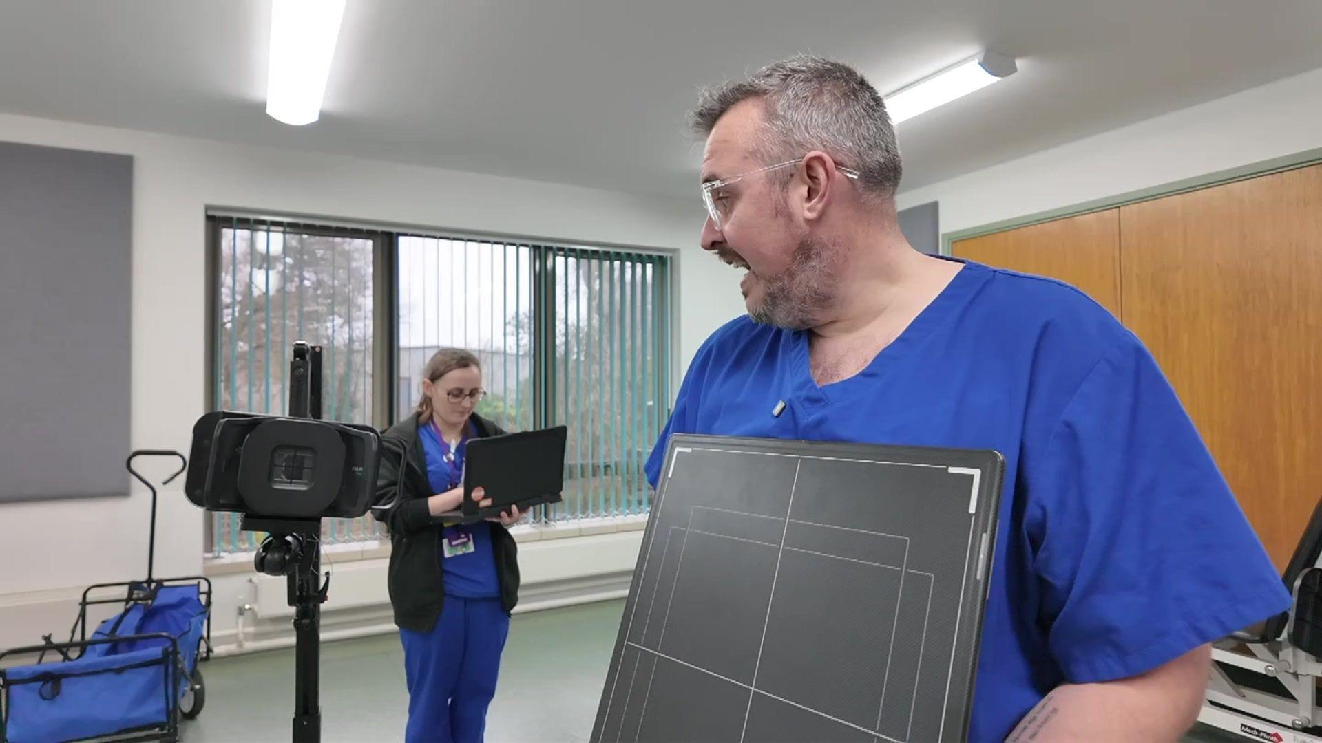 A mobile X-ray kit has been set up in a large grey room. A man and a woman are in the room wearing dark blue medical scrubs. The man, in the foreground, who has short grey hair, a beard, and glasses, is holding an X-ray imaging panel. In the background, a woman with long hair tied back is working on a laptop. An imaging device is mounted on a tripod in the room.