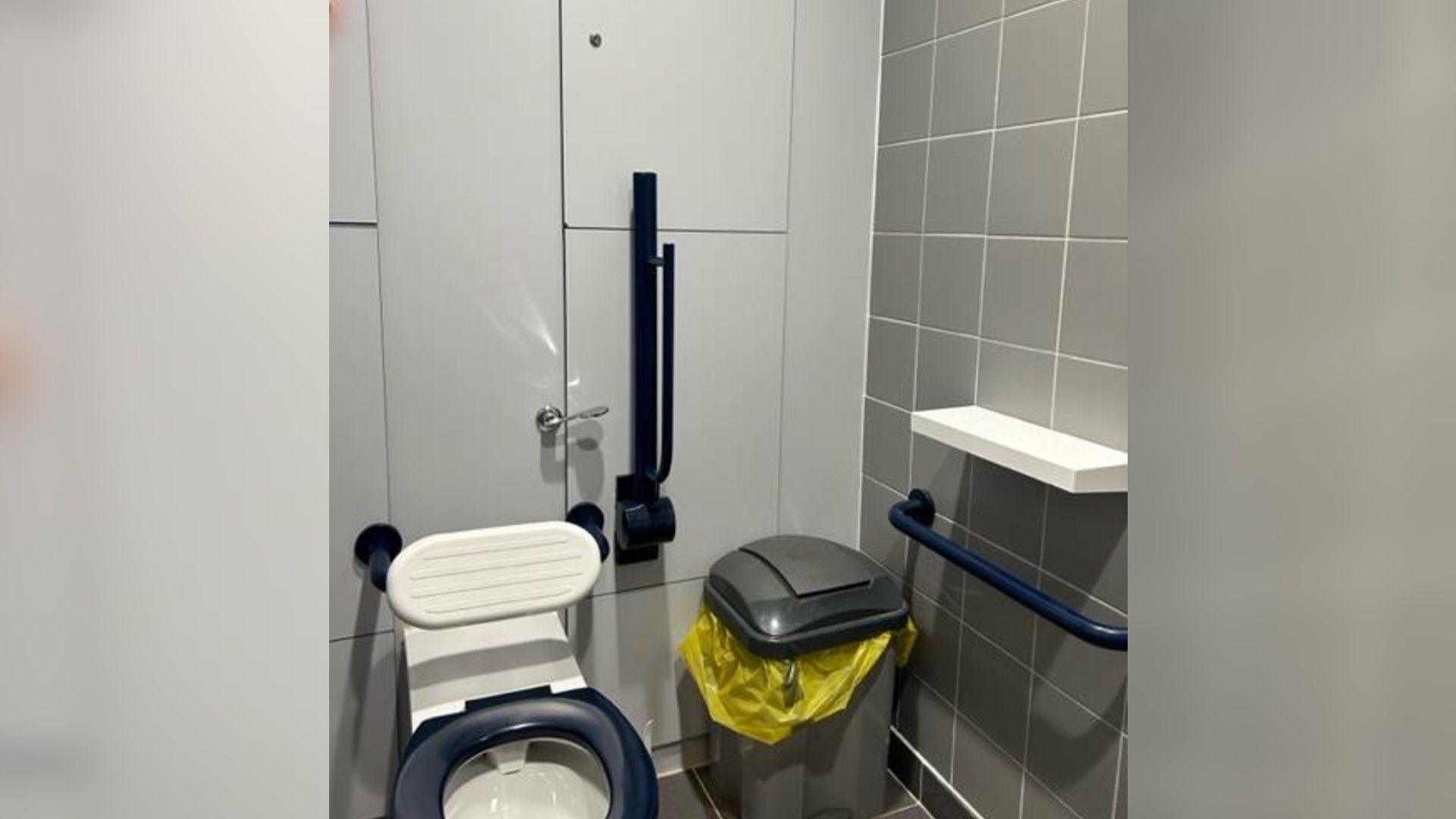 A toilet, a bin and a shelf in a railway station bathroom. The walls are white and grey.