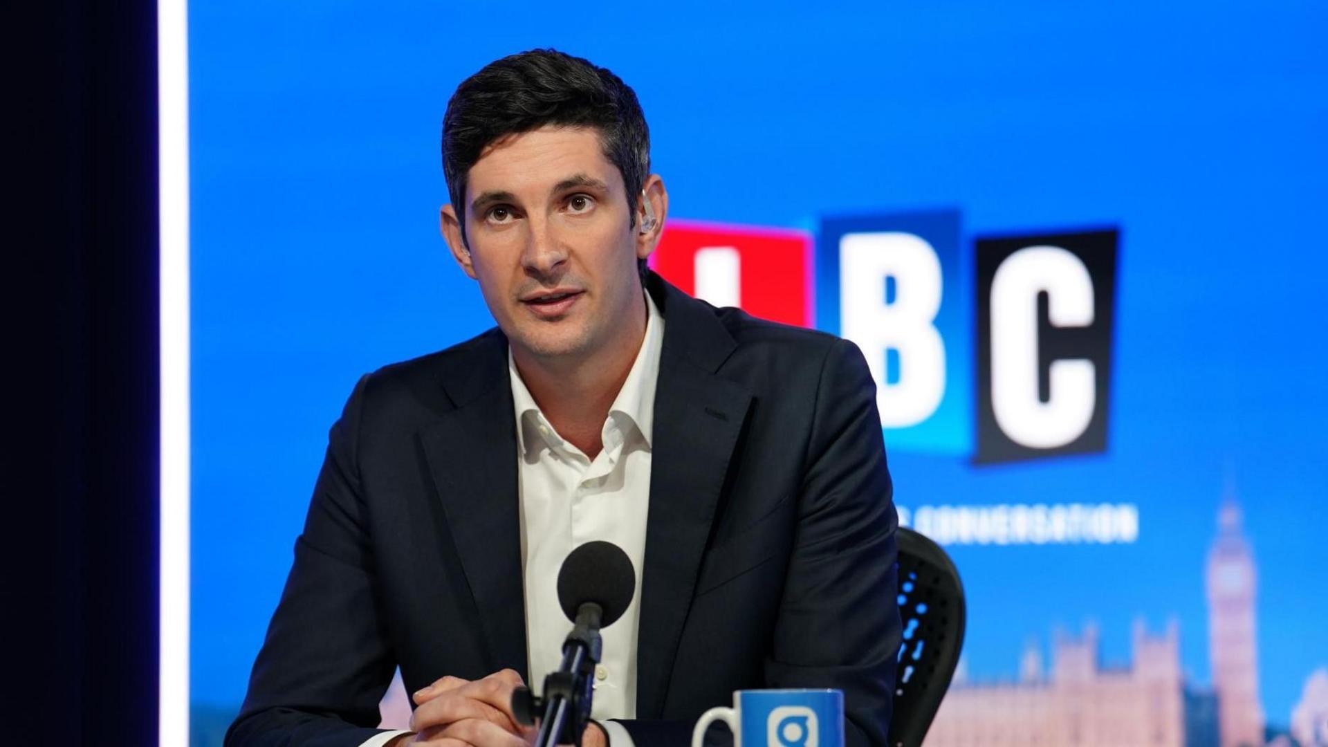 Tom Swarbrick in a navy suit jacket and white shirt, sat in front of a black microphone and with an earpiece in. In the background is a blue background with the outline of Westminster Palace visible and a red, blue and black logo that reads LBC.