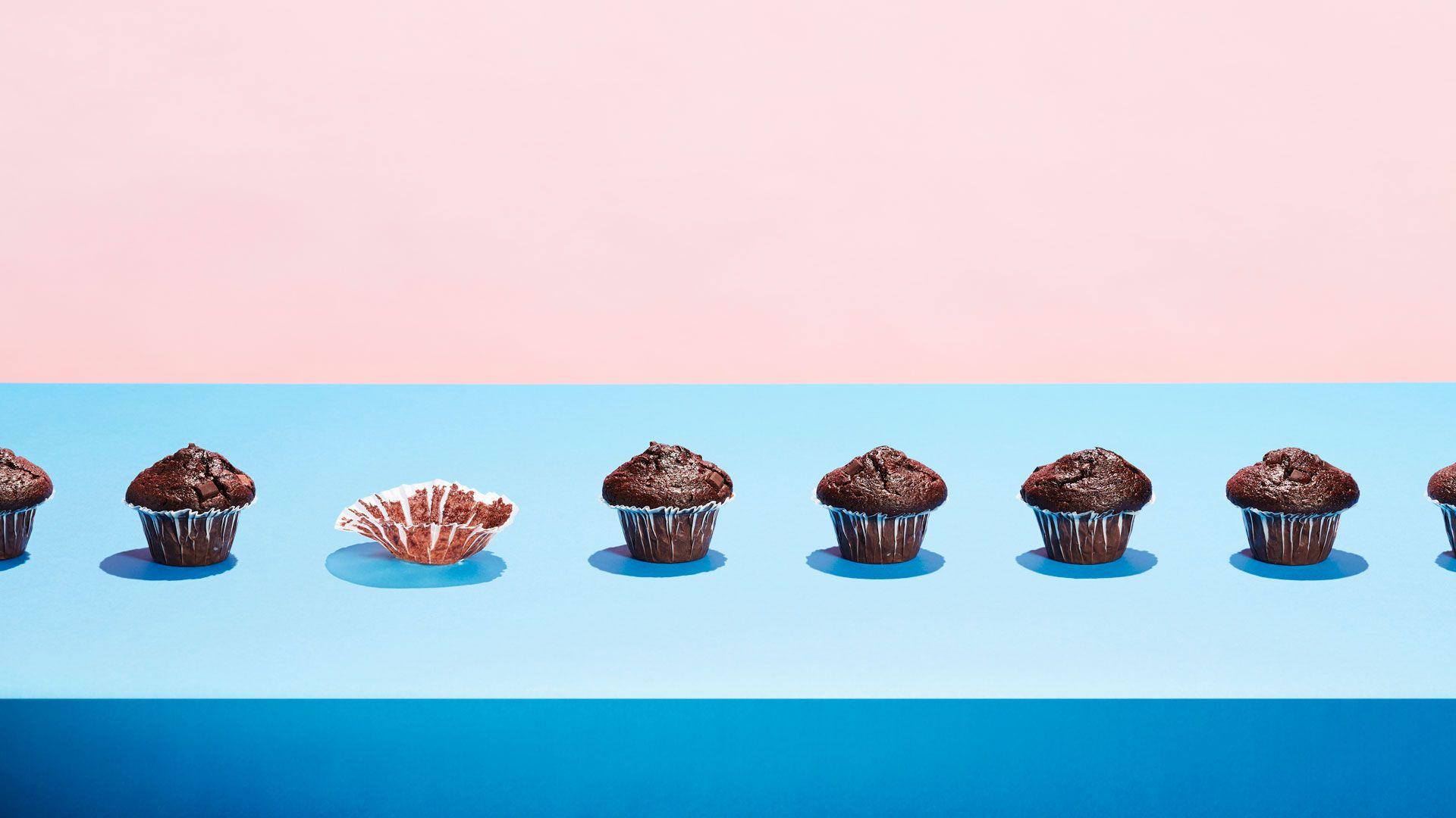 Chocolate muffins in a row with one eaten. On a pale blue and pink background