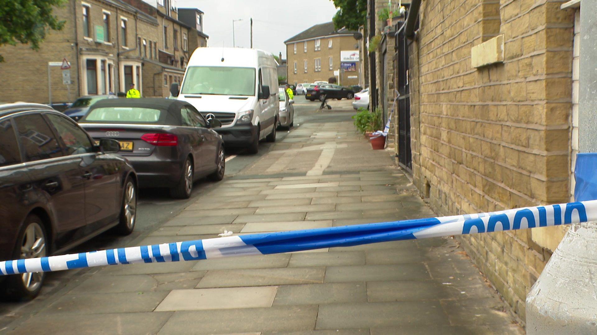 Police tape across a street in Bradford with terrace houses on each side, and two cars and a van parked by the pavement.