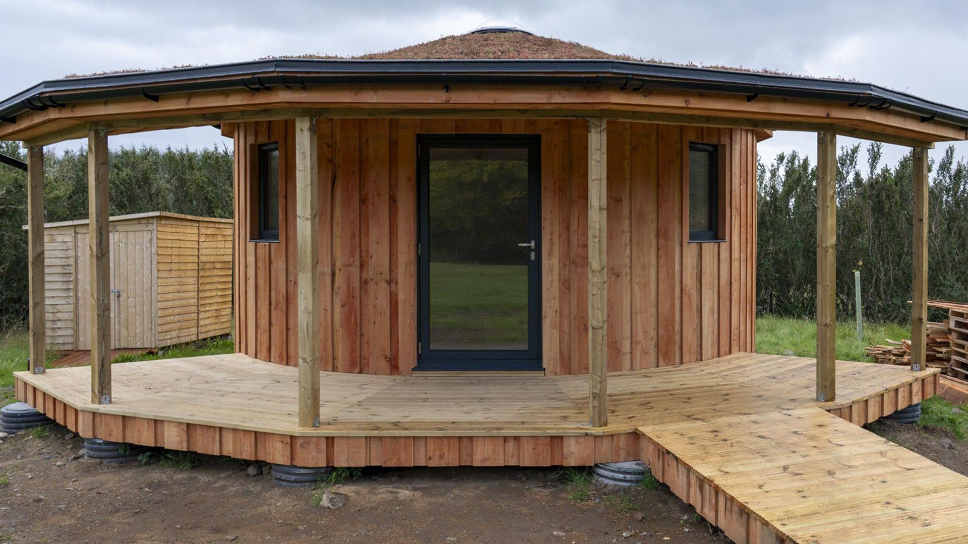 The centre has a round-shaped wooden building with a glass front door and windows in the walls. Around the building is a wooden, veranda-style structure. Trees surround the site.