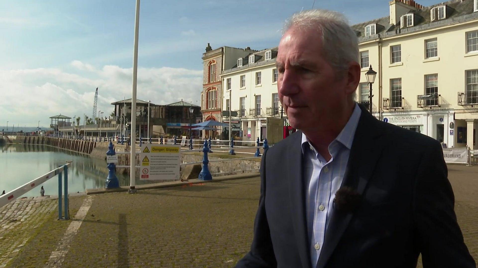 Councillor Chris Lewis. He is wearing a black suit with a light blue shirt and is standing in front of a harbour with buildings behind him on a cloudy day.