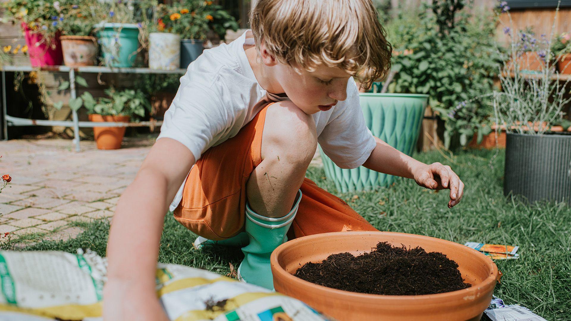 A boy wearing a white tshirt, orange shorts and teal wellies crouches on grass in a garden. Behind him in the background is a patio with pots of plants stacked on top of each other. There is a terracotta pot in front of him filled with soil. He is putting his hand into a bag of soil in the foreground. There are packets of seeds on the grass jut behind the rim of the pot. 