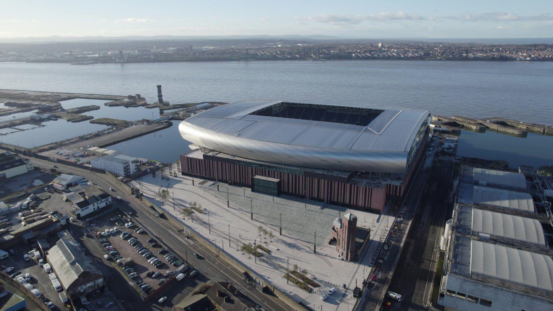 An aerial view of Everton's new stadium, a curved silver structure on dockland with the River Mersey in the backdrop.