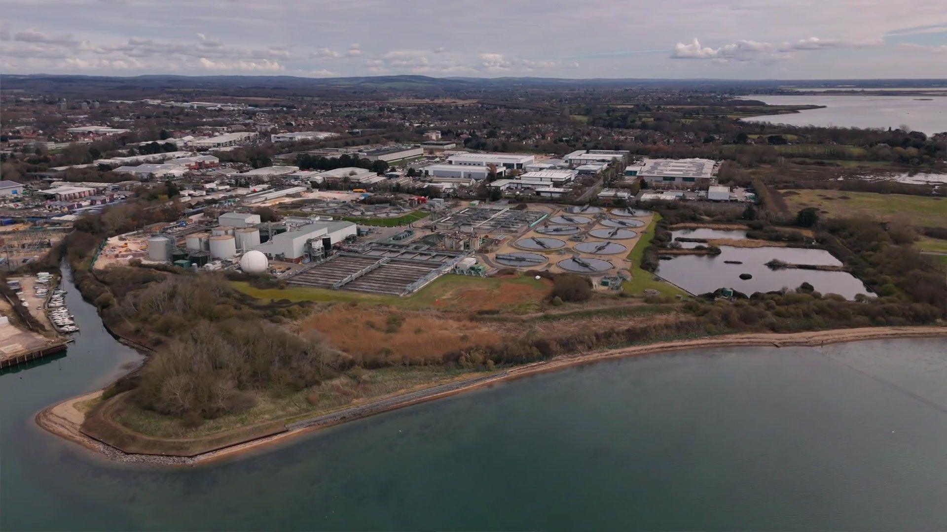 An aerial view of a water works in Portsmouth, can see a section of water which is surrounded by trees and bushes, large pillar buildings and industrial plants.