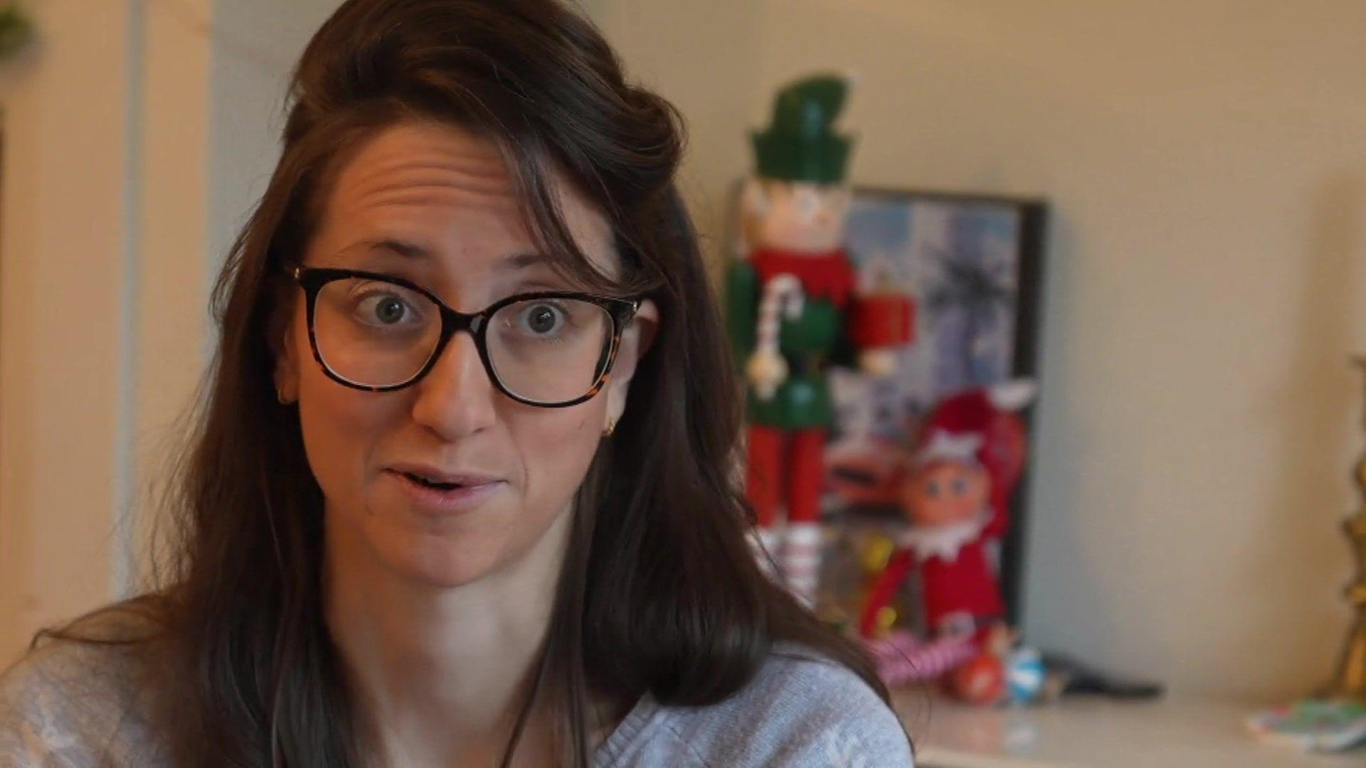 Katie, who has long straight brown hair and wears black-rimmed glasses, speaks to a BBC reporter. Behind her are elf Christmas decorations in her living room