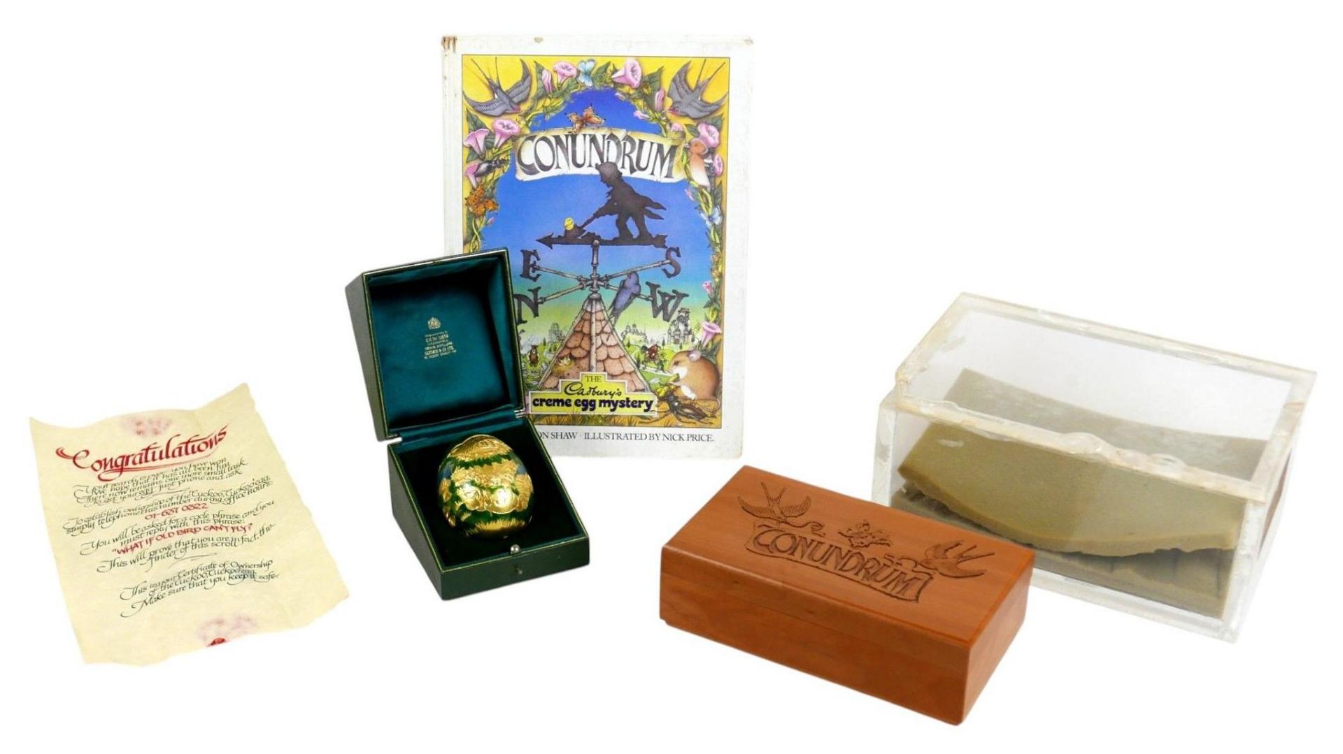 A photo of a congratulations letter, a Conundrum illustrated magazine, the golden egg, a wooden box, and a Perspex box, all taken on a white background.