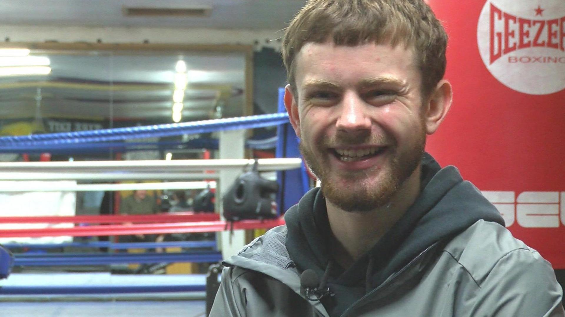 Reuben smiling at the camera, he has hazel facial hair and is wearing a grey jacket over a black hoodie. In the background is the boxing ring. 