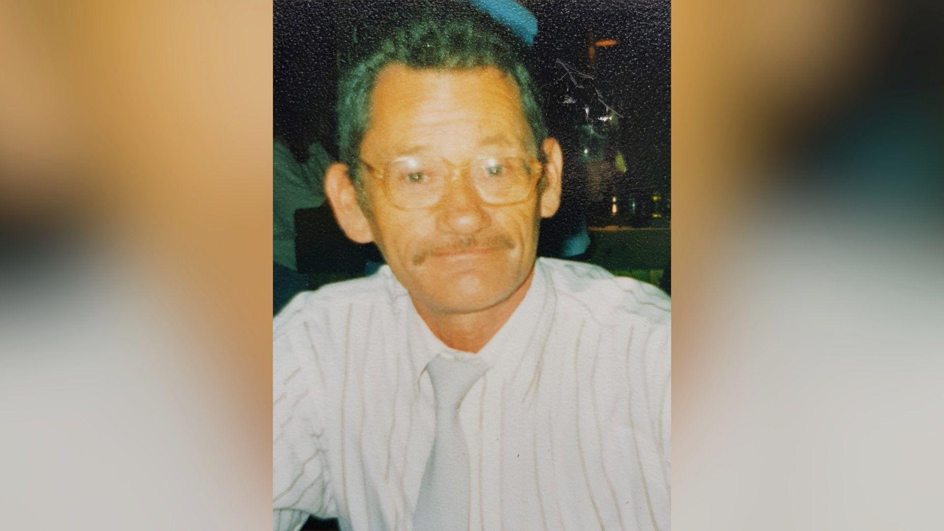 A man in his fifties, with short brown hair and glasses is looking at the camera. He has a moustache and is smiling. It is an old photograph and some of the colour has drained from it. He is wearing a shirt and tie.