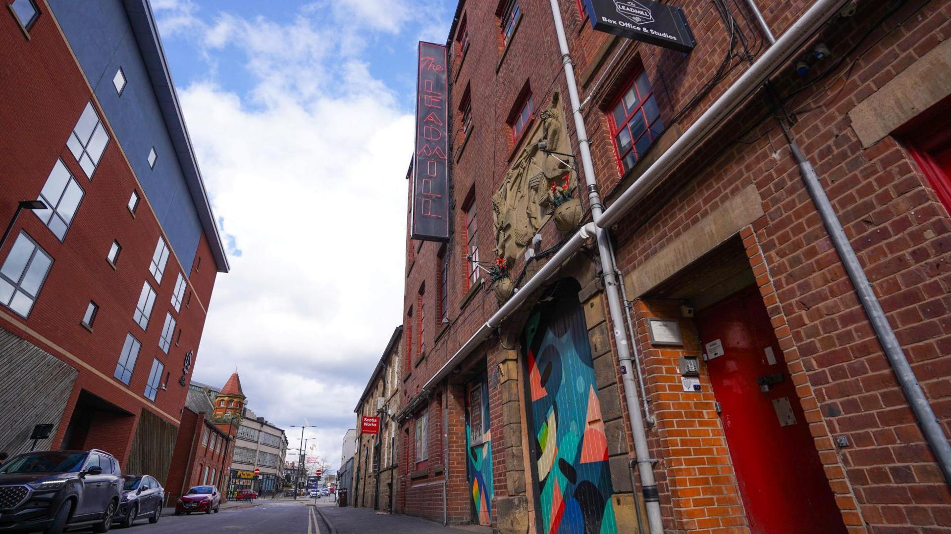 A street with a four-storey building  on one side. There is a black sign outside with the words 'The Leadmill' in red, neon writing. Two doors to the building are painted in multi-colours. 