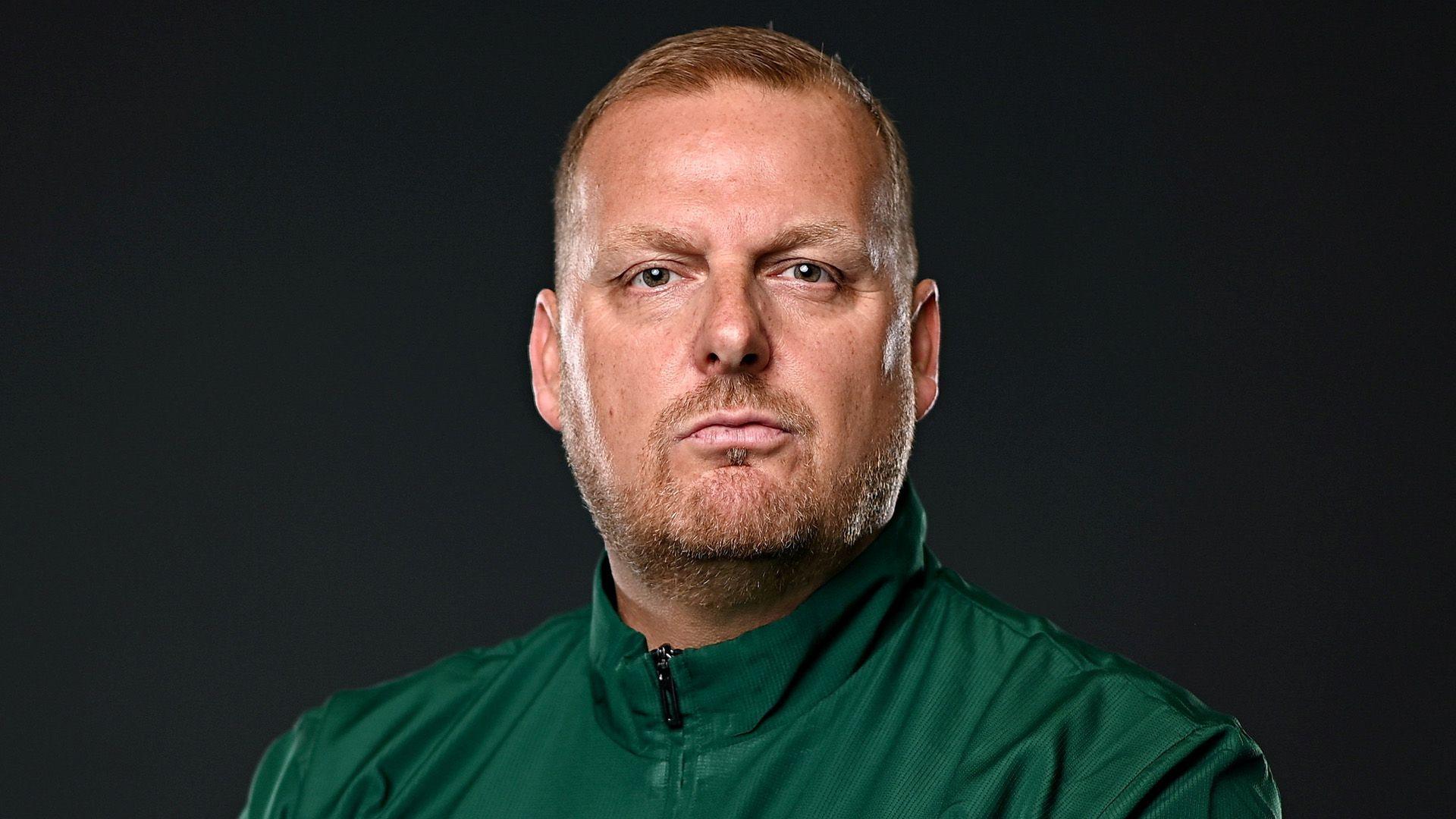 Headshot of Jon Rudd who is looking straight at the camera with a serious expression on his face. He has short fair hair and beard and is wearing a dark green tracksuit top. He is pictured against a black background.