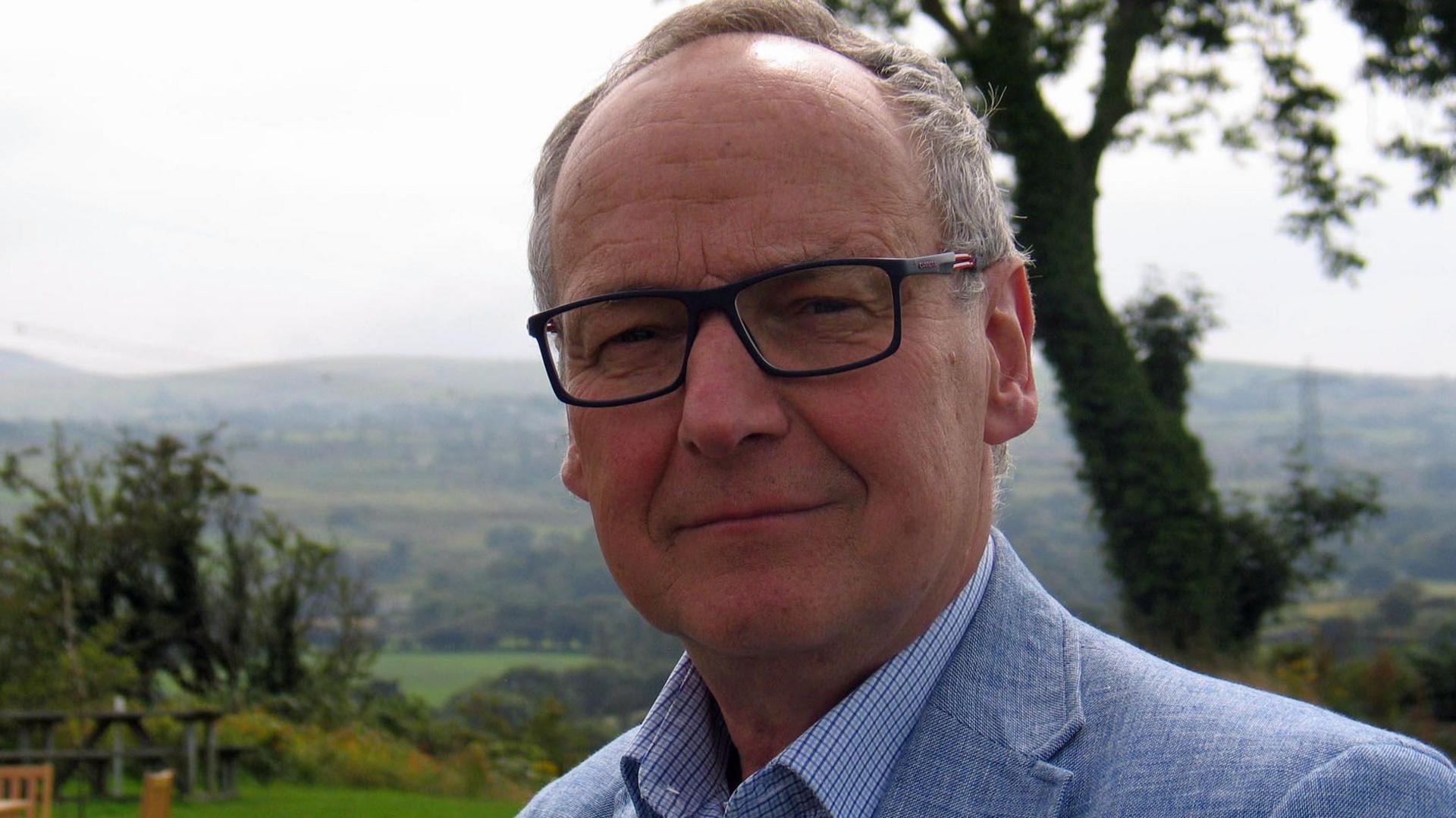 Dyfrig Siencyn standing in a green field with hills and trees in the background. He has glasses on and is wearing a blue jacket and blue shirt