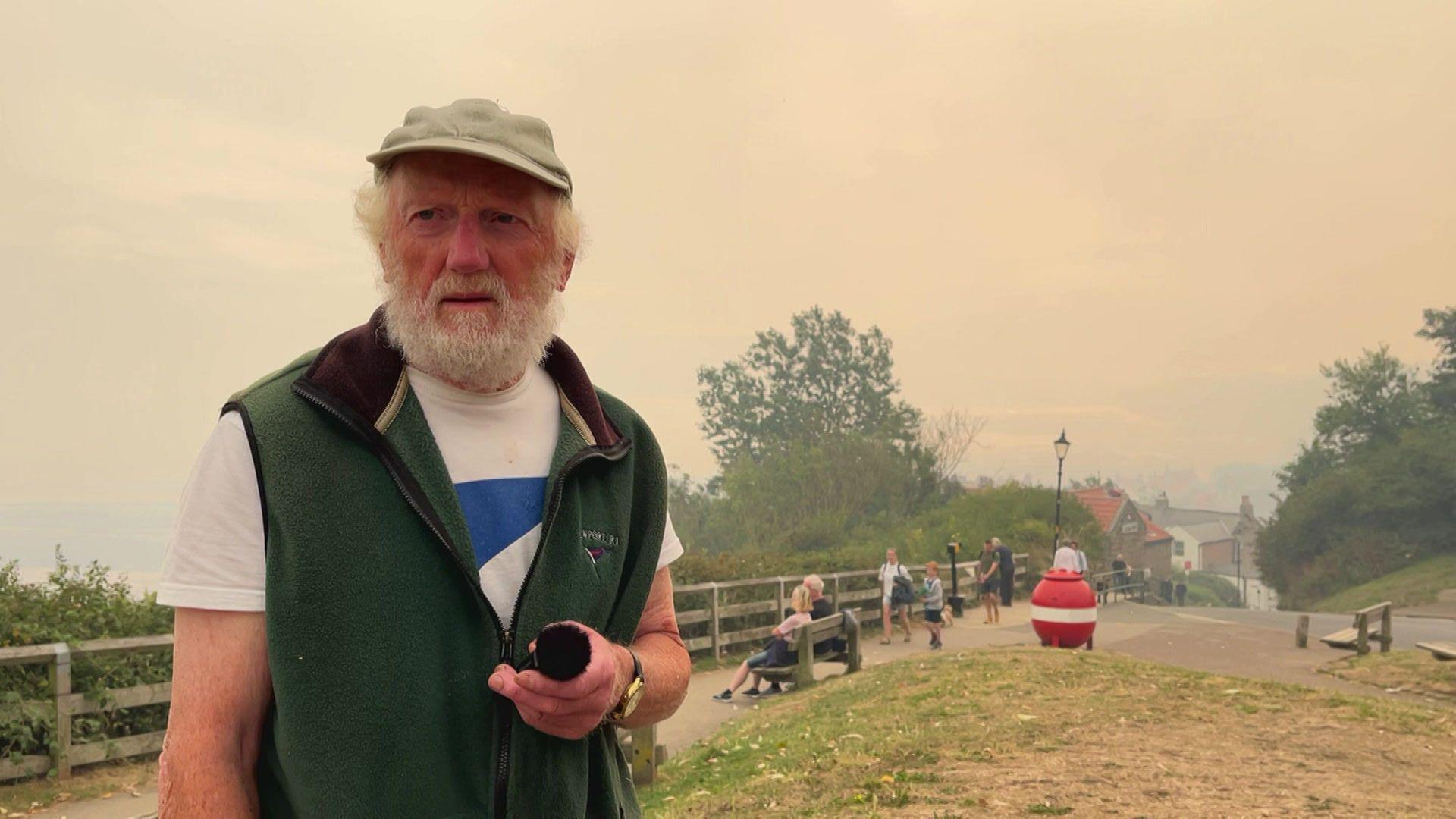 An older, grey-bearded man, wearing a green gilet, light T-shirt with a blue pattern on the chest, and green cap stands in front of a backdrop of Robin Hood's Bay. The air is visibly smoky.