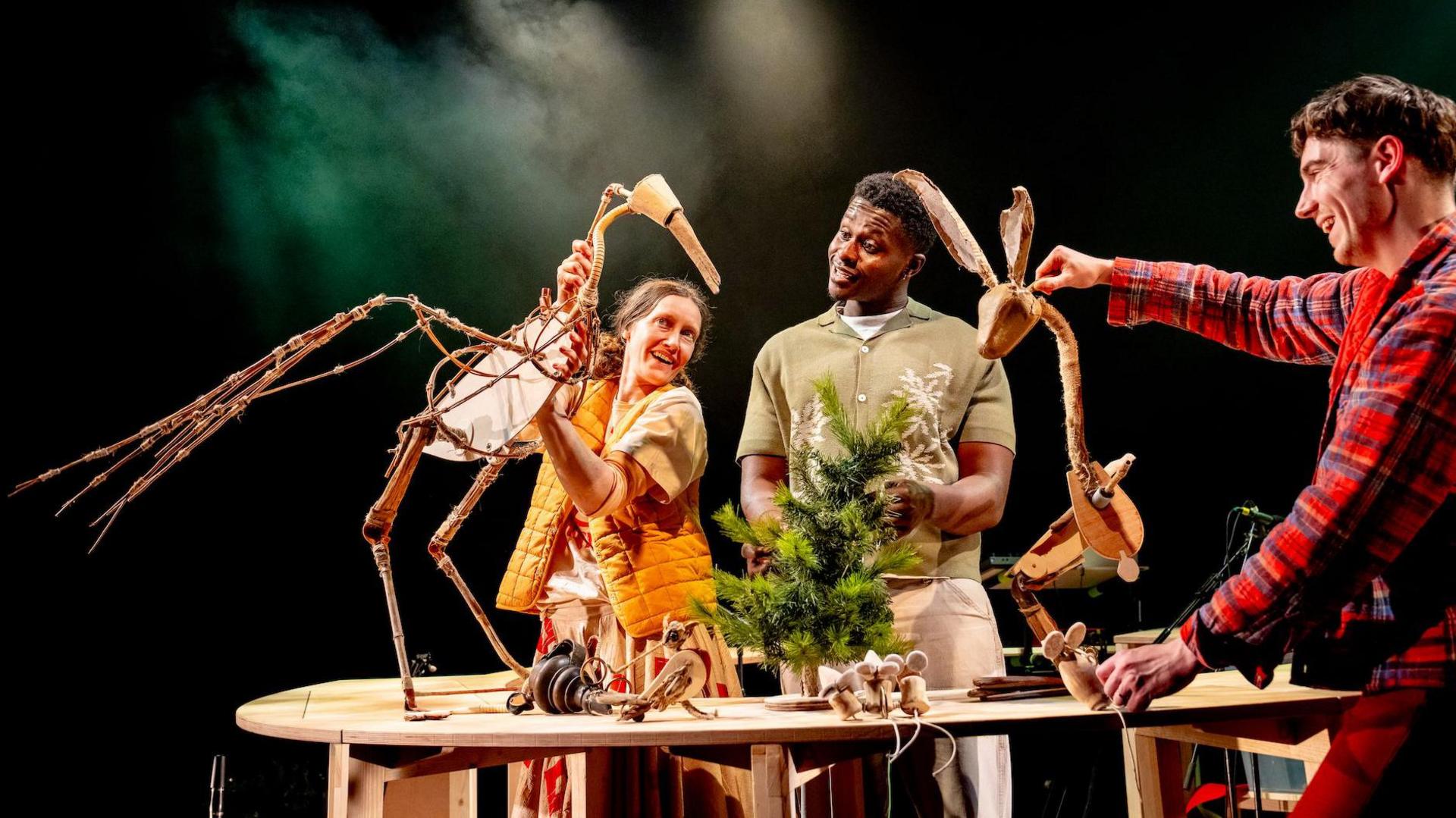 Three people on stage under lights. One holds a puppet that looks like a heron bird, another a puppet that looks like a hare. A tiny fir tree is in the middle of the picture