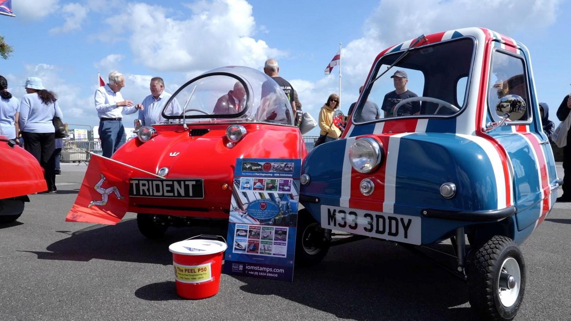 A P50, painted in the red, white and blue of the union jack flag is parked up next to a red Trident car, which has a Manx flag attached to the front. People are standing behind them admiring them.