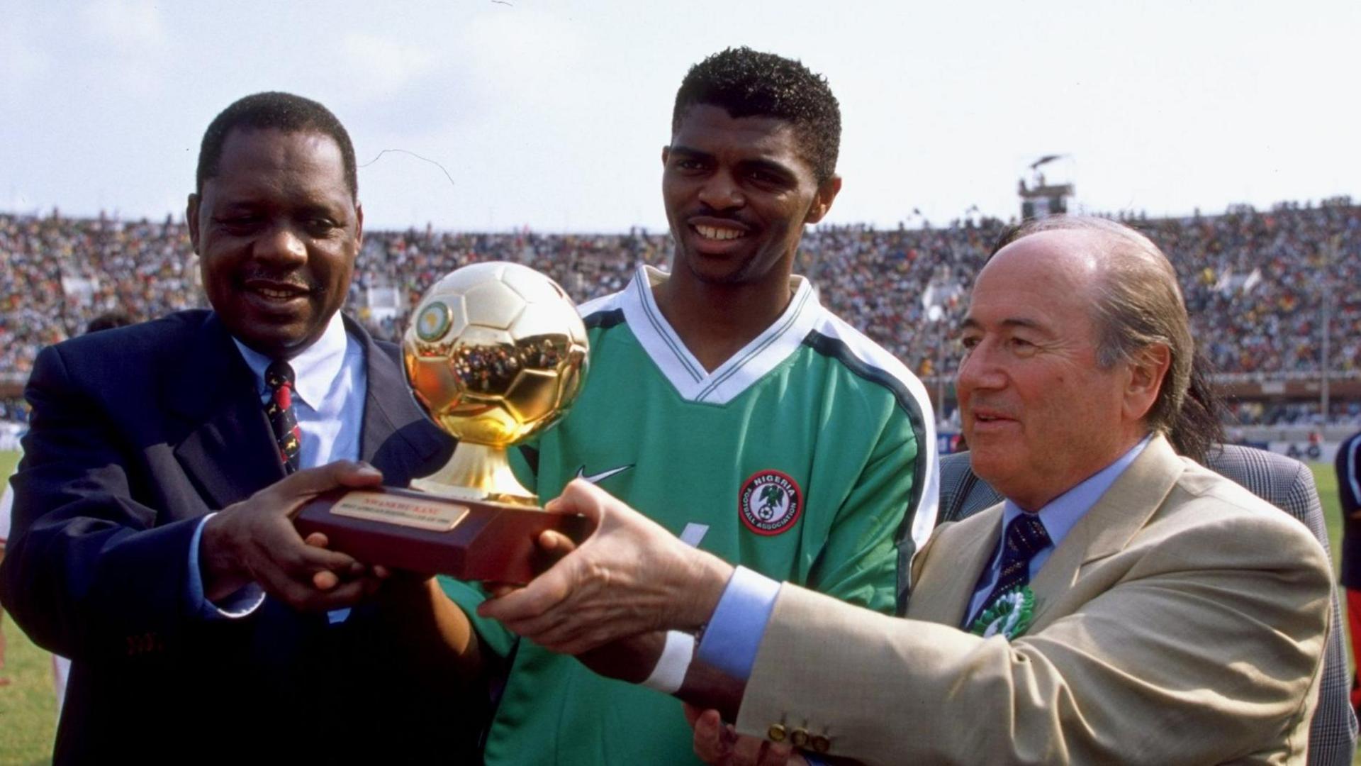 Issa Hayatou, in a dark blue suit, and Sepp Blatter, in a cream suit, hand a golden ball trophy for the African Footballer of the Year award to Nwankwo Kanu, who is wearing a green Nigeria shirt. Full stands in an open-air football stadium can be seen in the background