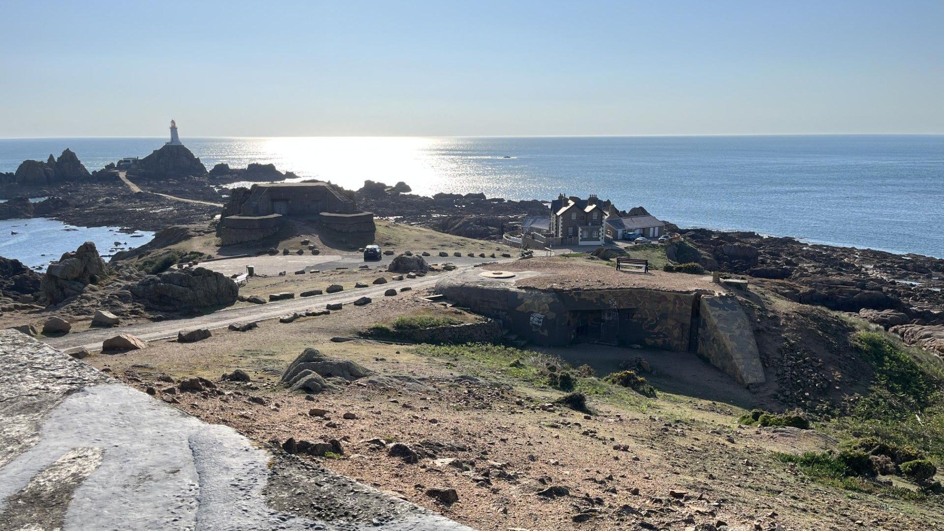 A picture of La Corbière. The lighthouse can be seen in the background to the left. There is a bunker on a hill and a car parked outside it. To the right there are houses. The sea is in the background and the sun is lighting the sea up. The sky is blue.